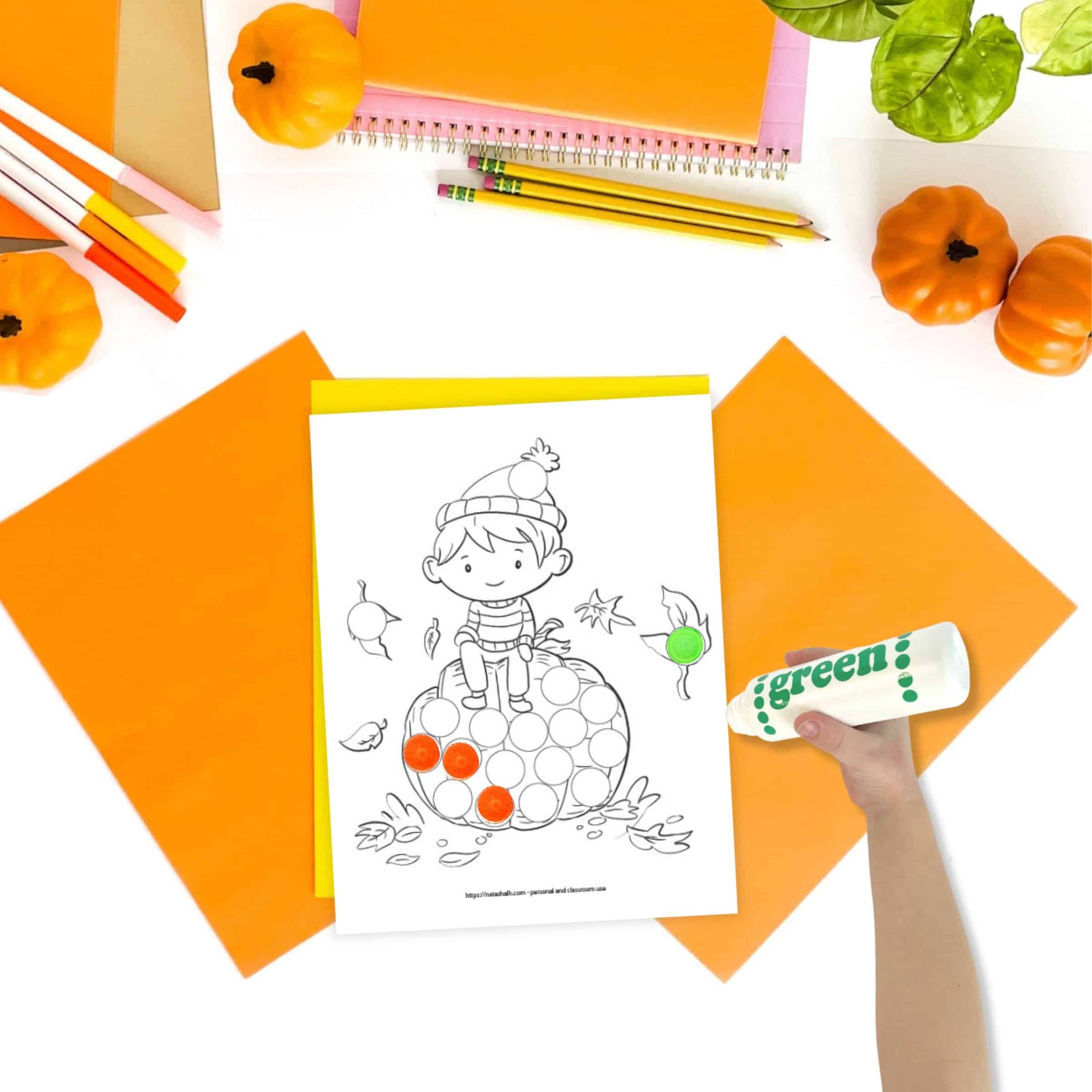 A top down image of a dot marker coloring page showing a boy sitting on a large pumpkin. The page is shown on a desk with orange paper and small pumpkins