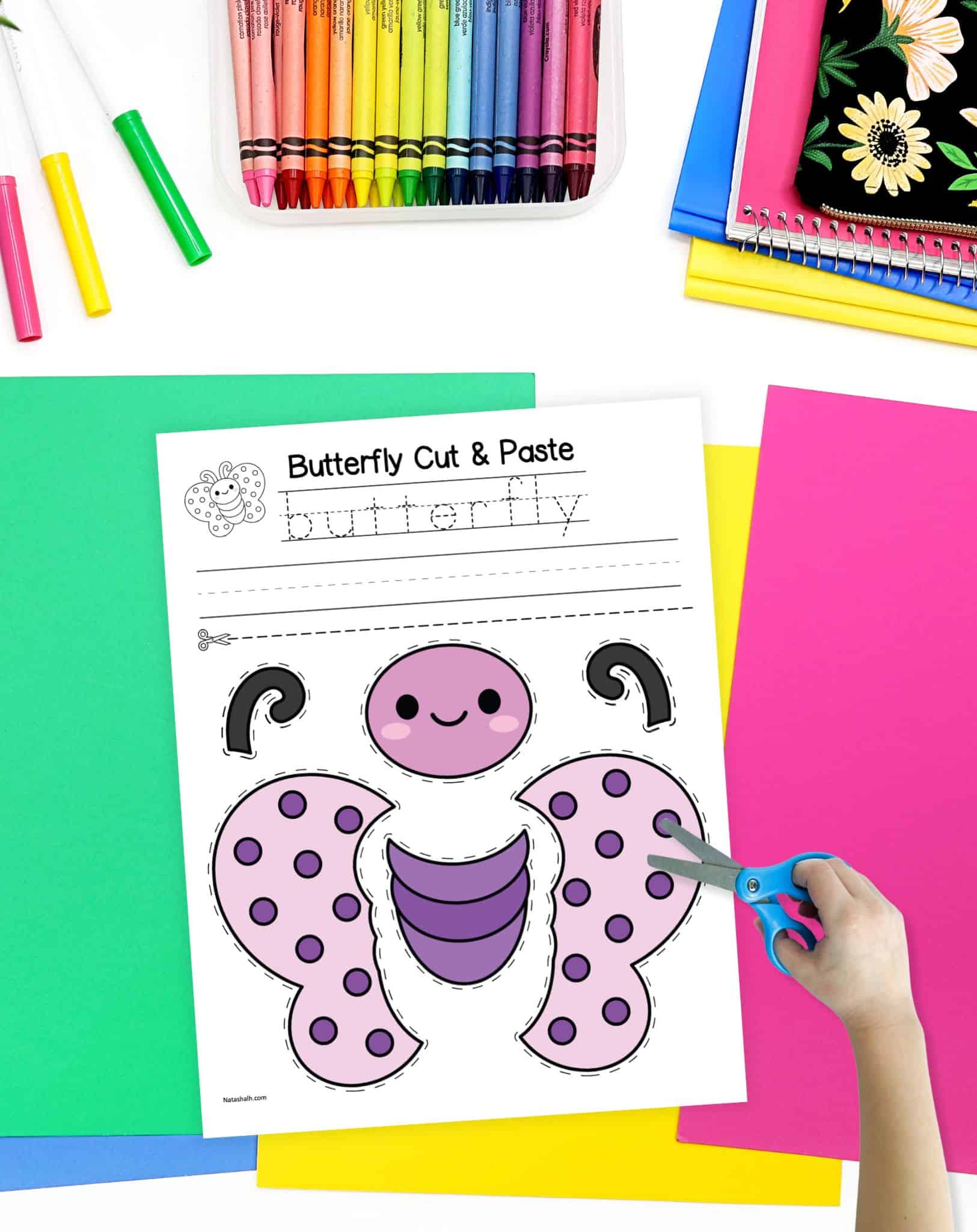 A top down image of a desk with colorful papers, pencils, and a cut and paste butterfly craft. A child's hand holding a pair of scissors is available in the foreground.