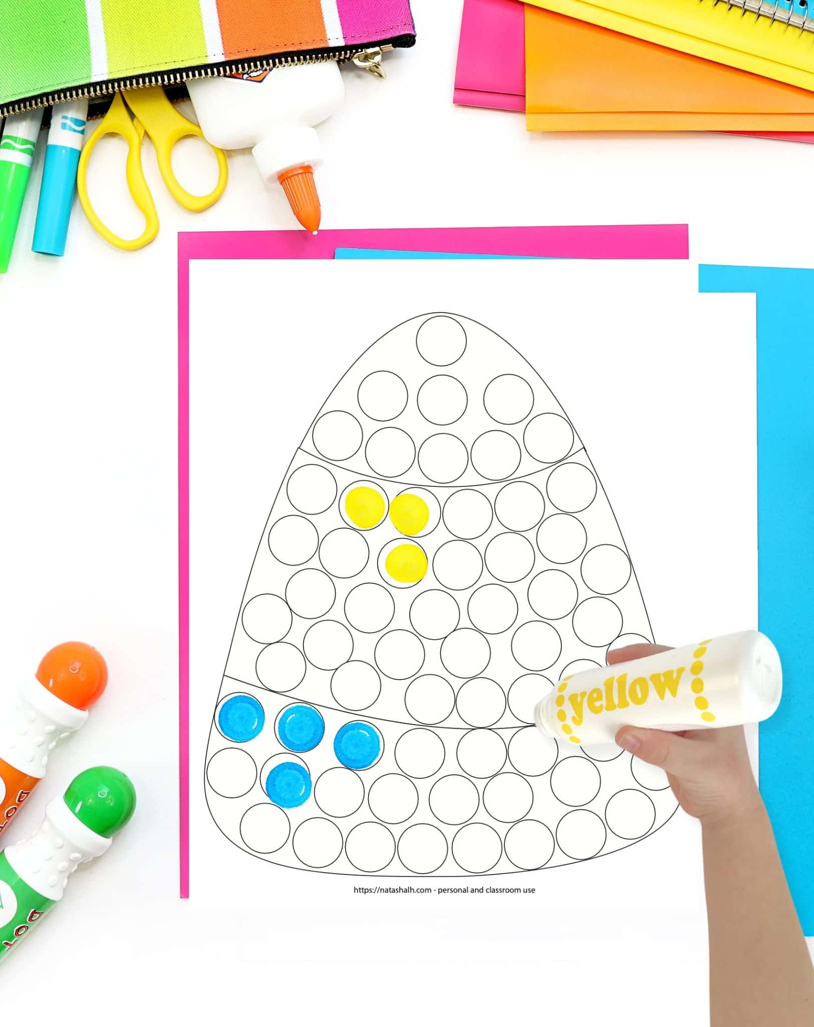 A top down image of a candy corn themed dot marker coloring page. It is on a table with colorful papers and a child's hand holding a yellow dot marker is visible in the foreground