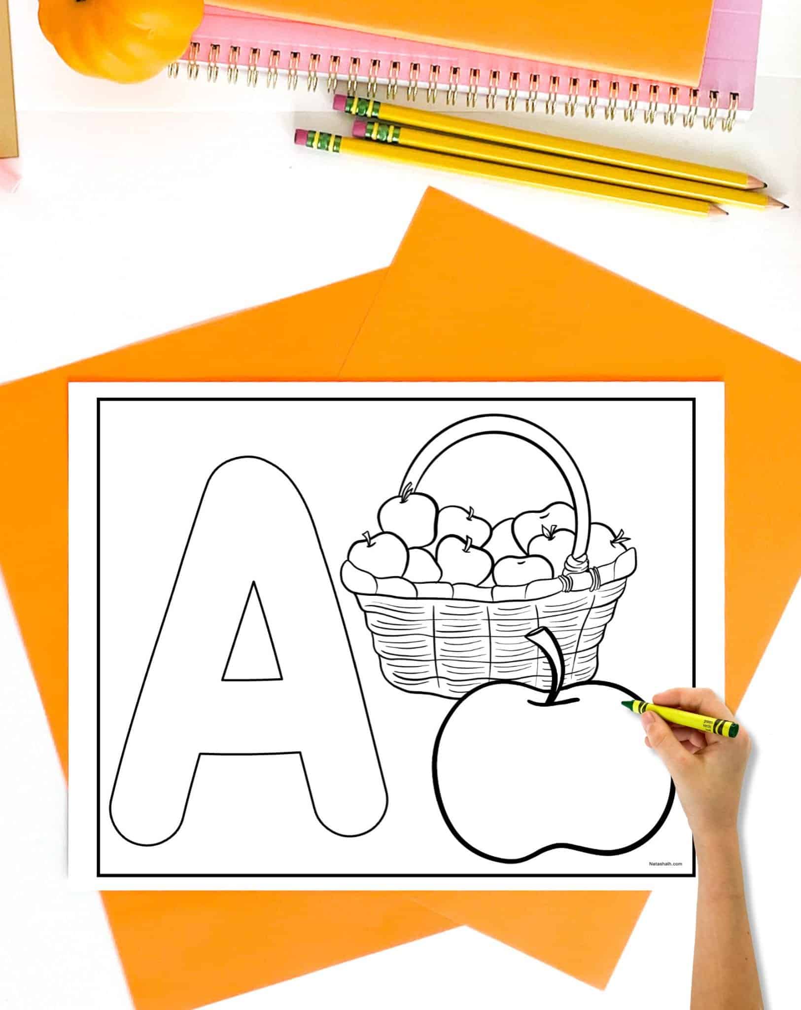 An uppercase bubble a to color with a basket of apples and a single apple in front of the basket. The page is on a desk with orange papers. A child's hand holding a crayon is visible in the foreground.