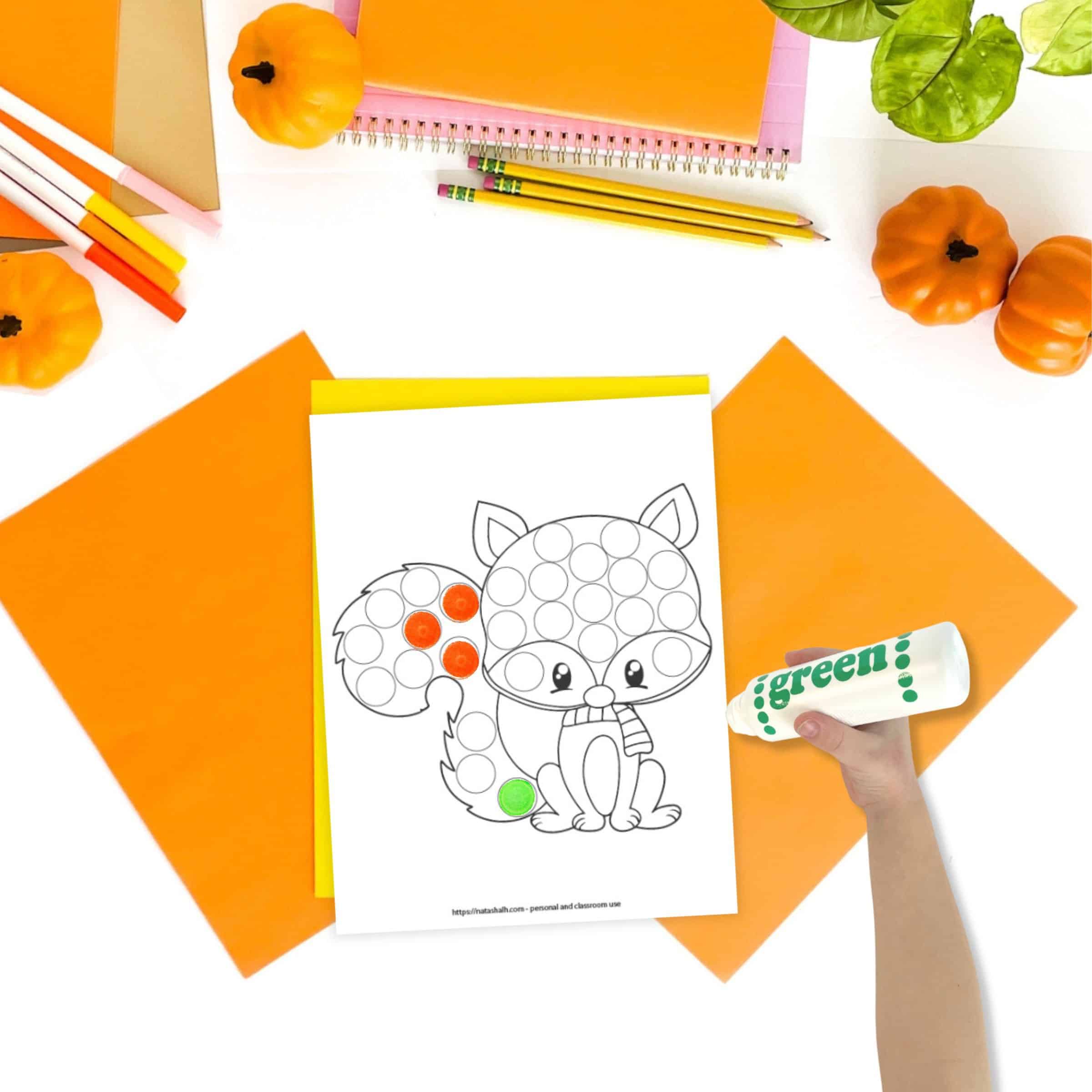 A top down image of a fox themed dot marker coloring page on a desk with orange papers and small pumpkins