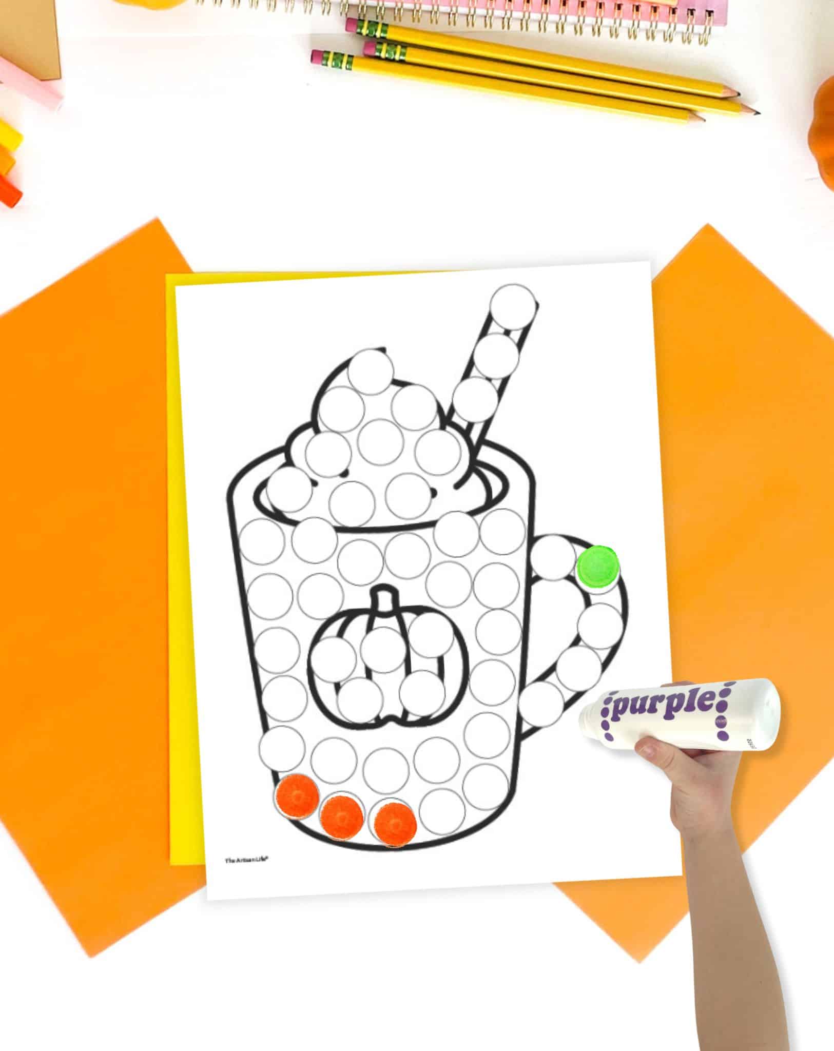 A dot marker page showing a cup of hot chocolate with whipped cream. It is on a table with fall colored papers. A hand holding a dot marker is visible in the foreground.