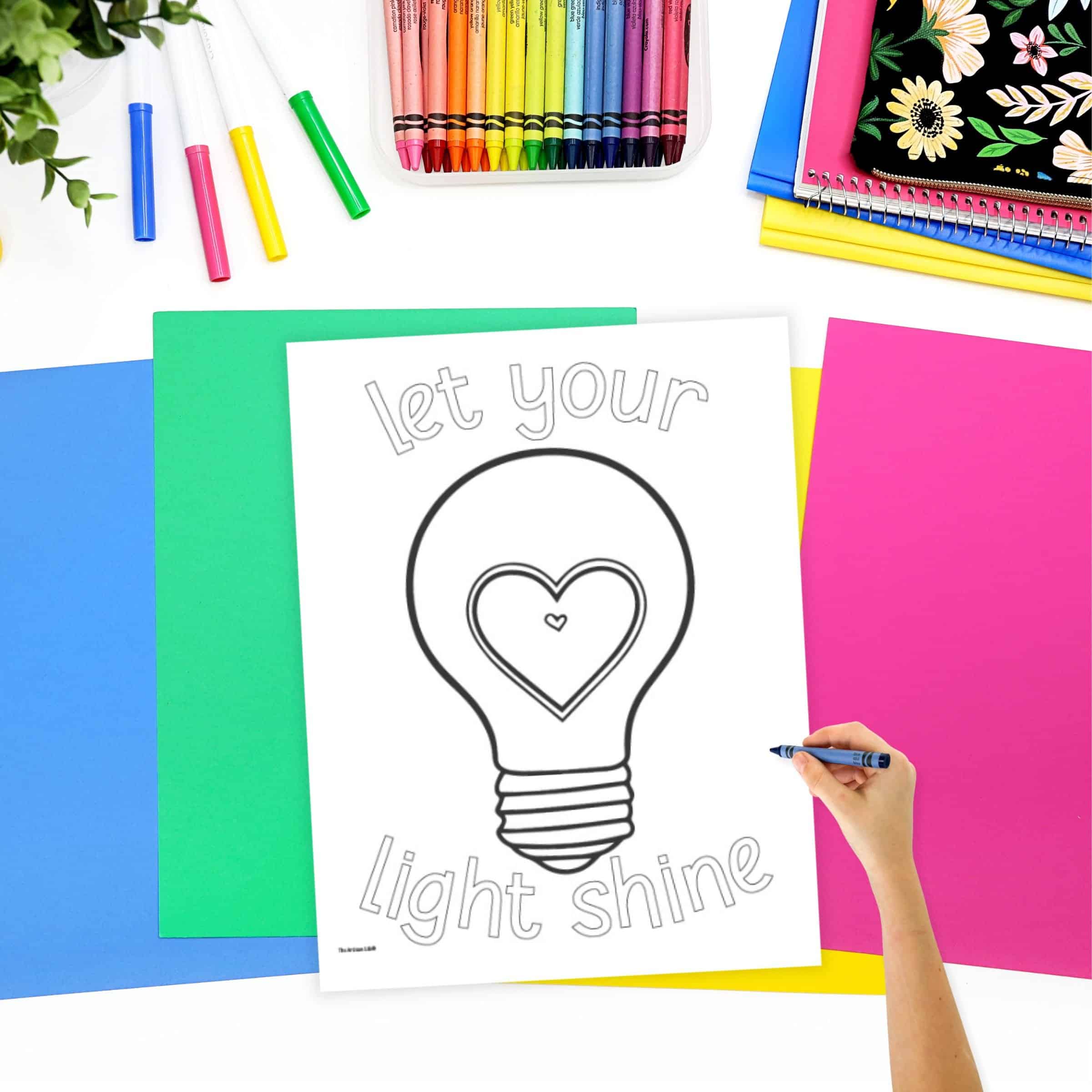 A light bulb coloring page on a desk with colorful pages. A child's hand holding a crayon is visible in the foreground