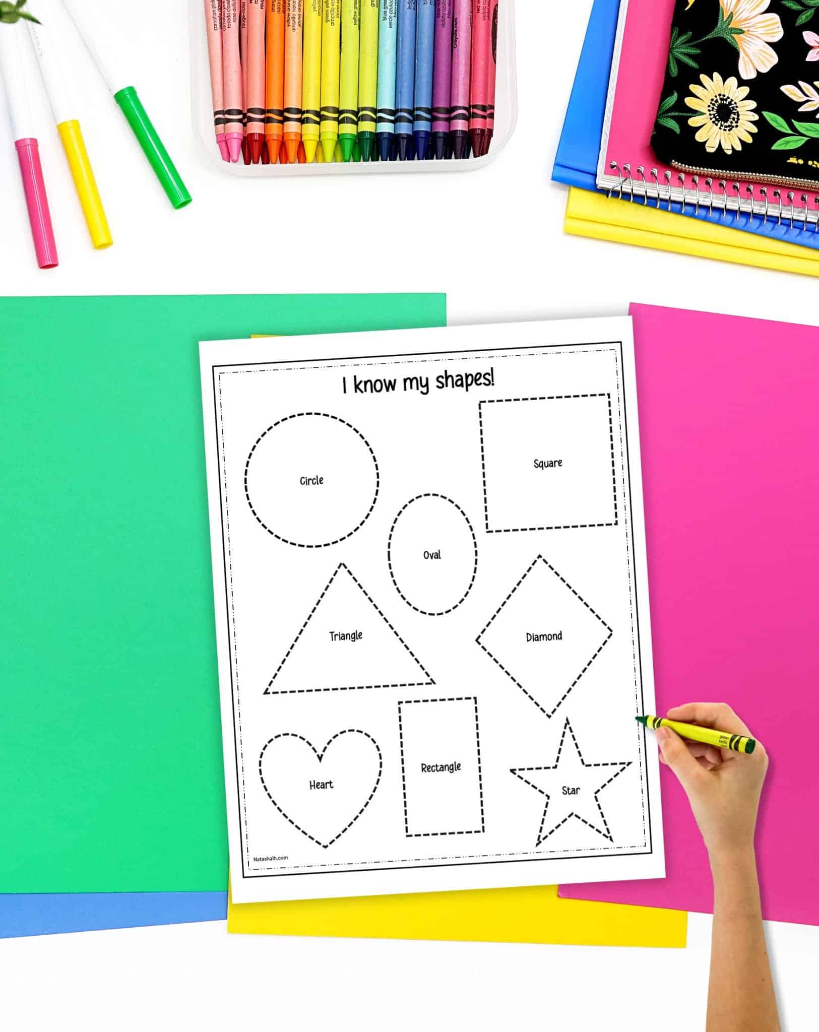 A shape tracing worksheet on a desk with colorful papers and crayons. A child's hand holding a green crayon is visible in the foreground.