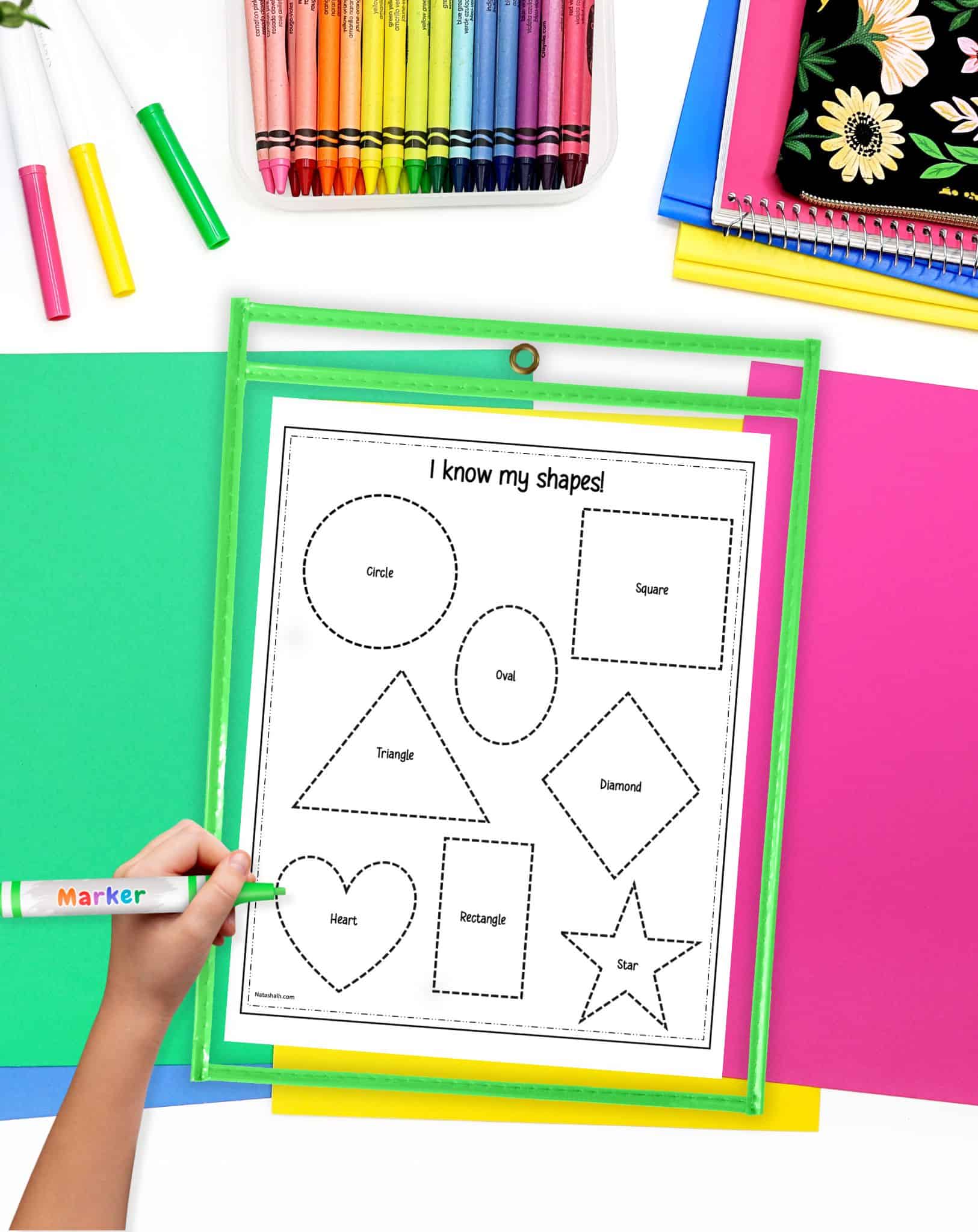 A shape tracing worksheet on a desk with colorful papers and crayons. The worksheet is in ad ry erase pocket and there is a child's hand holding a green marker