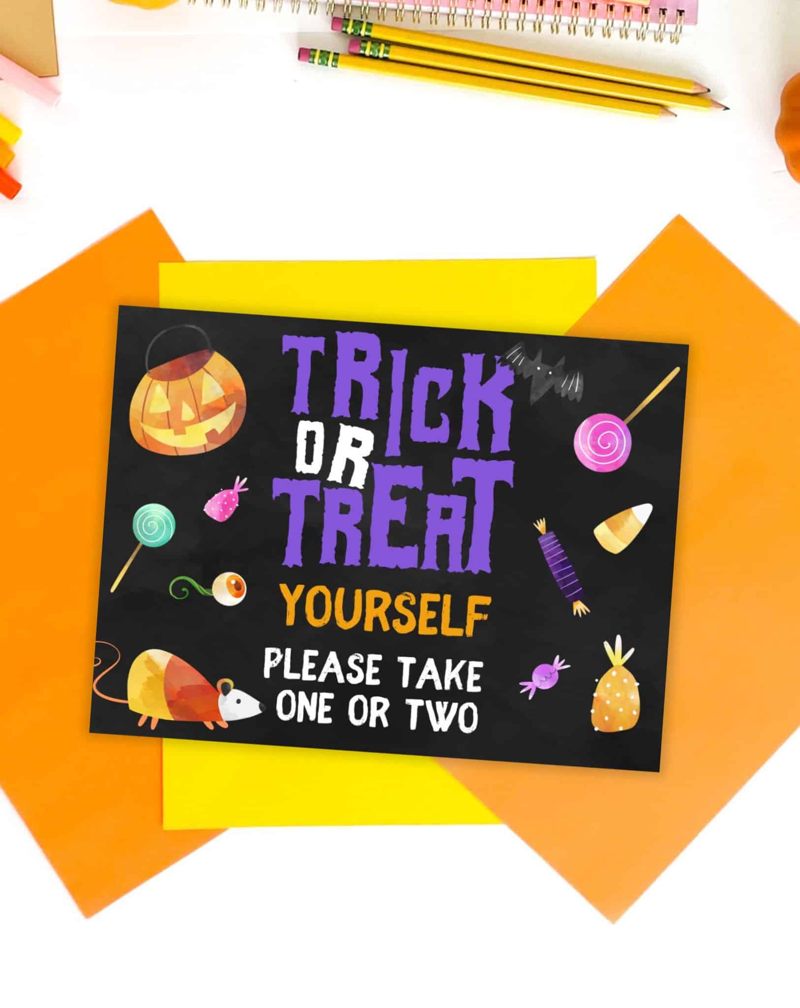 A top down photo of Halloween colored papers and pumpkins with a trick or treat sign printable. The sign has a chalkboard background and asks guests to take one or two pieces of candy.