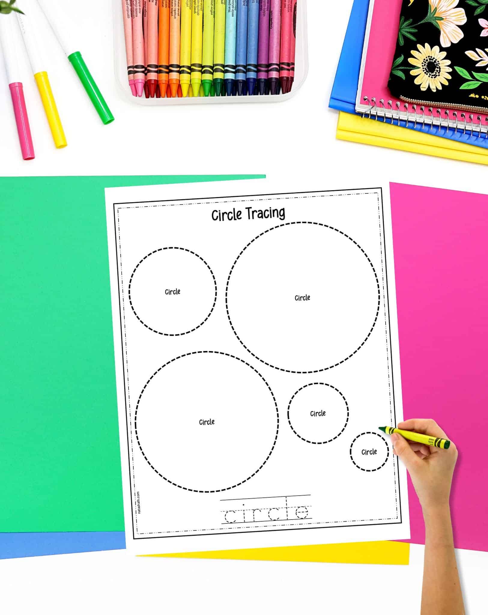 A circle tracing worksheet on a desk with colorful papers and crayons. A child's hand holding a crayon is visible in the foreground.