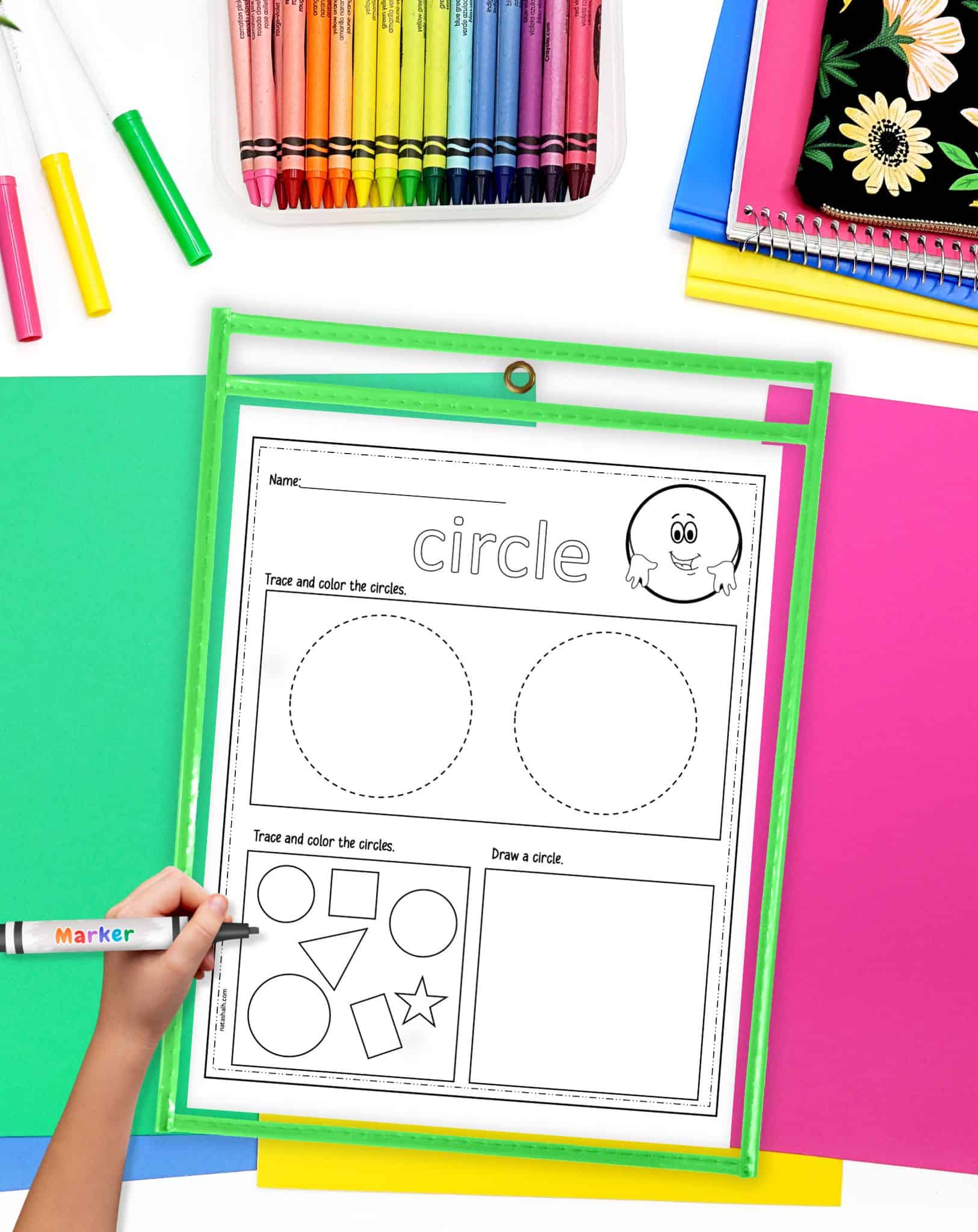 A circle themed worksheet in a dry erase pocket on a desk with colorful papers and crayons. A child's hand holding a marker is visible in the foreground.