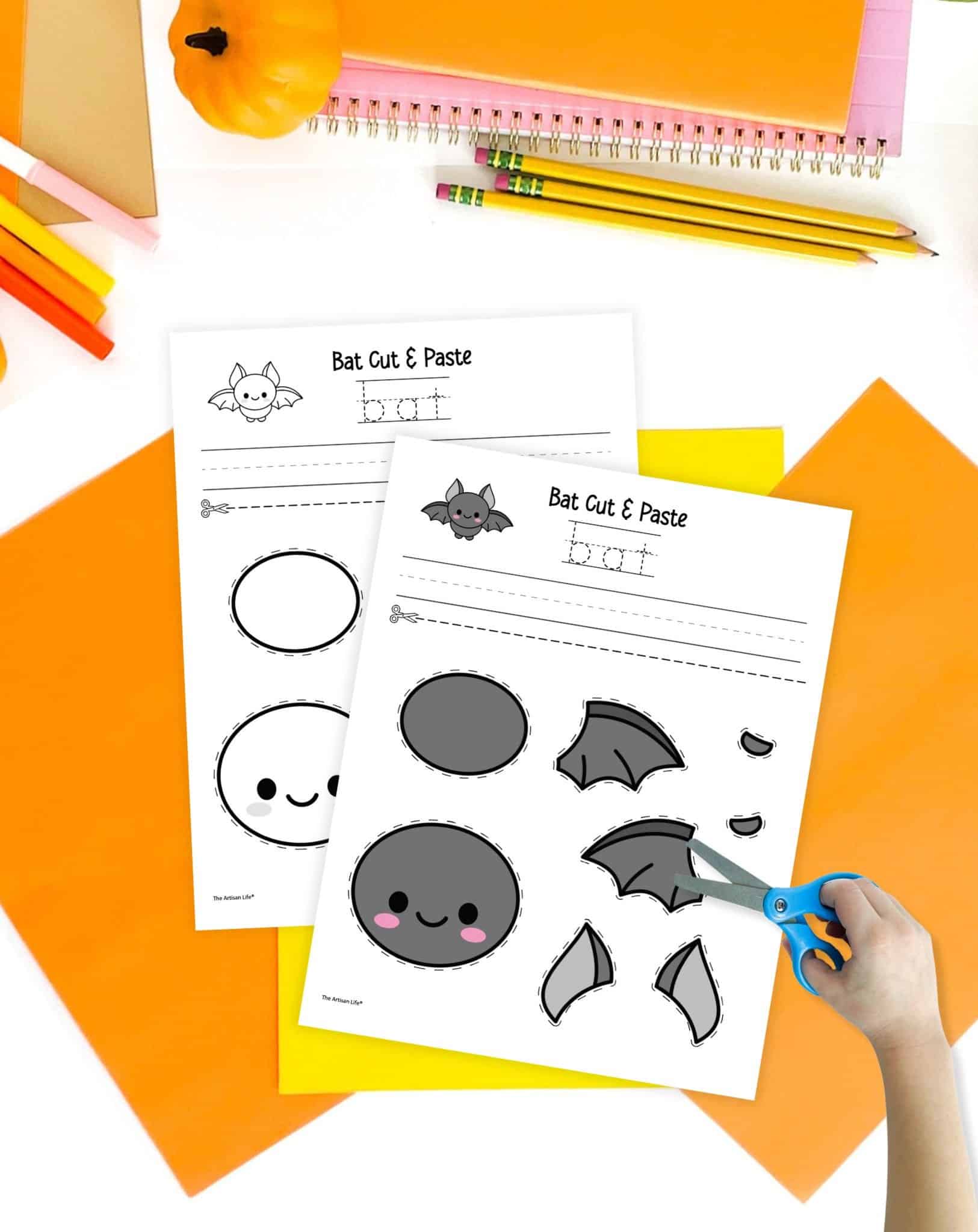 Two bat craft printables on a table with orange and yellow papers. A child's hand holding scissors is visible in the foreground.