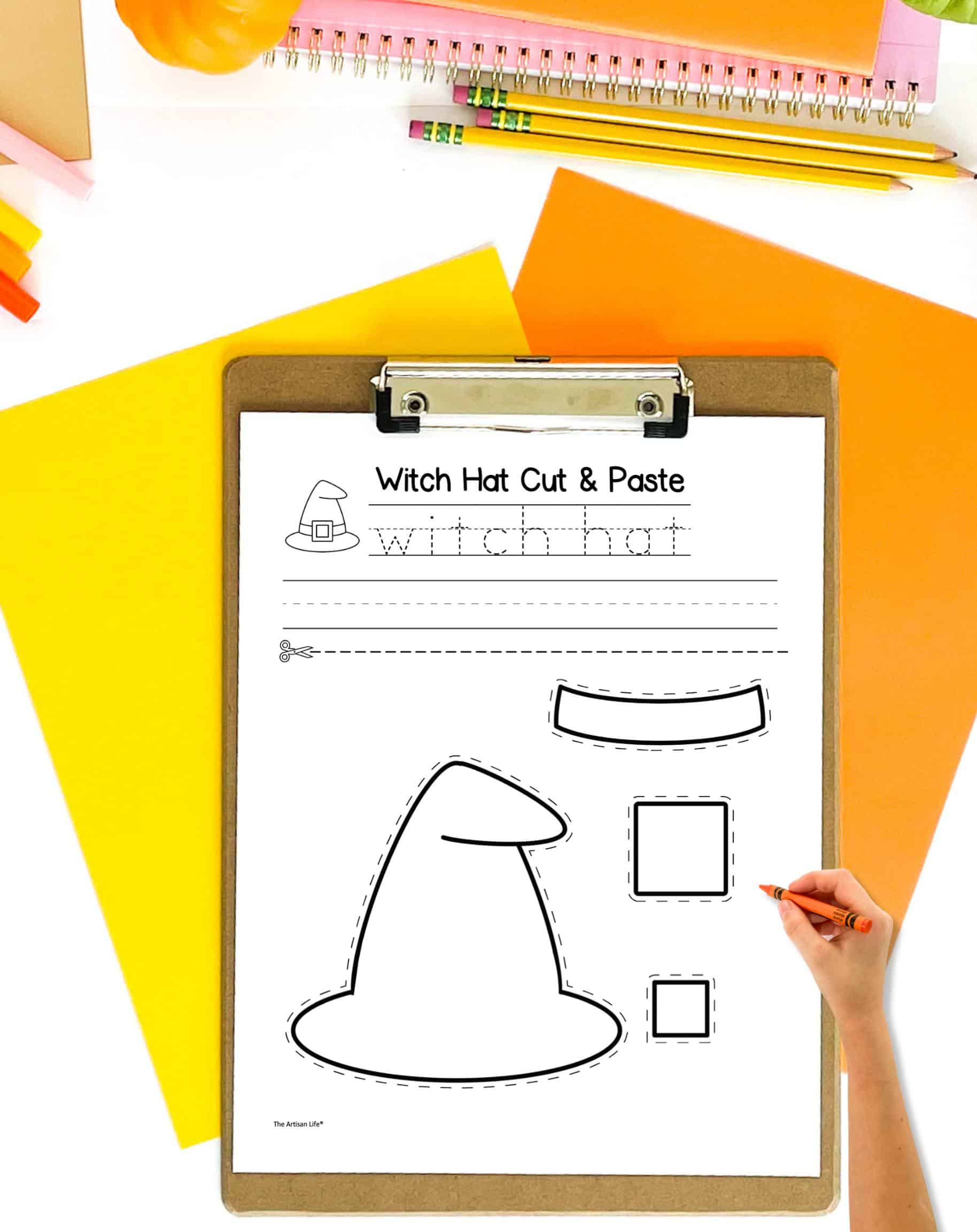 A witch hat cut and paste craft with handwriting practice. It is on a clip board. The page is on a desk with orange and yellow papers. A child's hand holding a crayon is visible in the foreground.