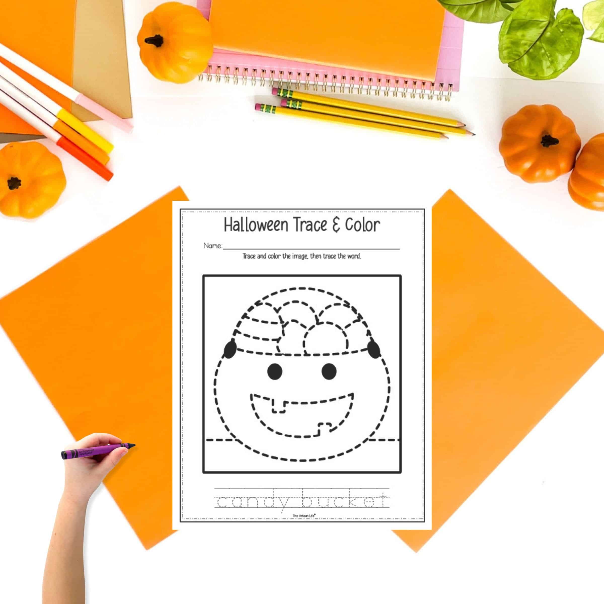 A Halloween trace and color page showing a candy bucket on a table with orange papers and pencils