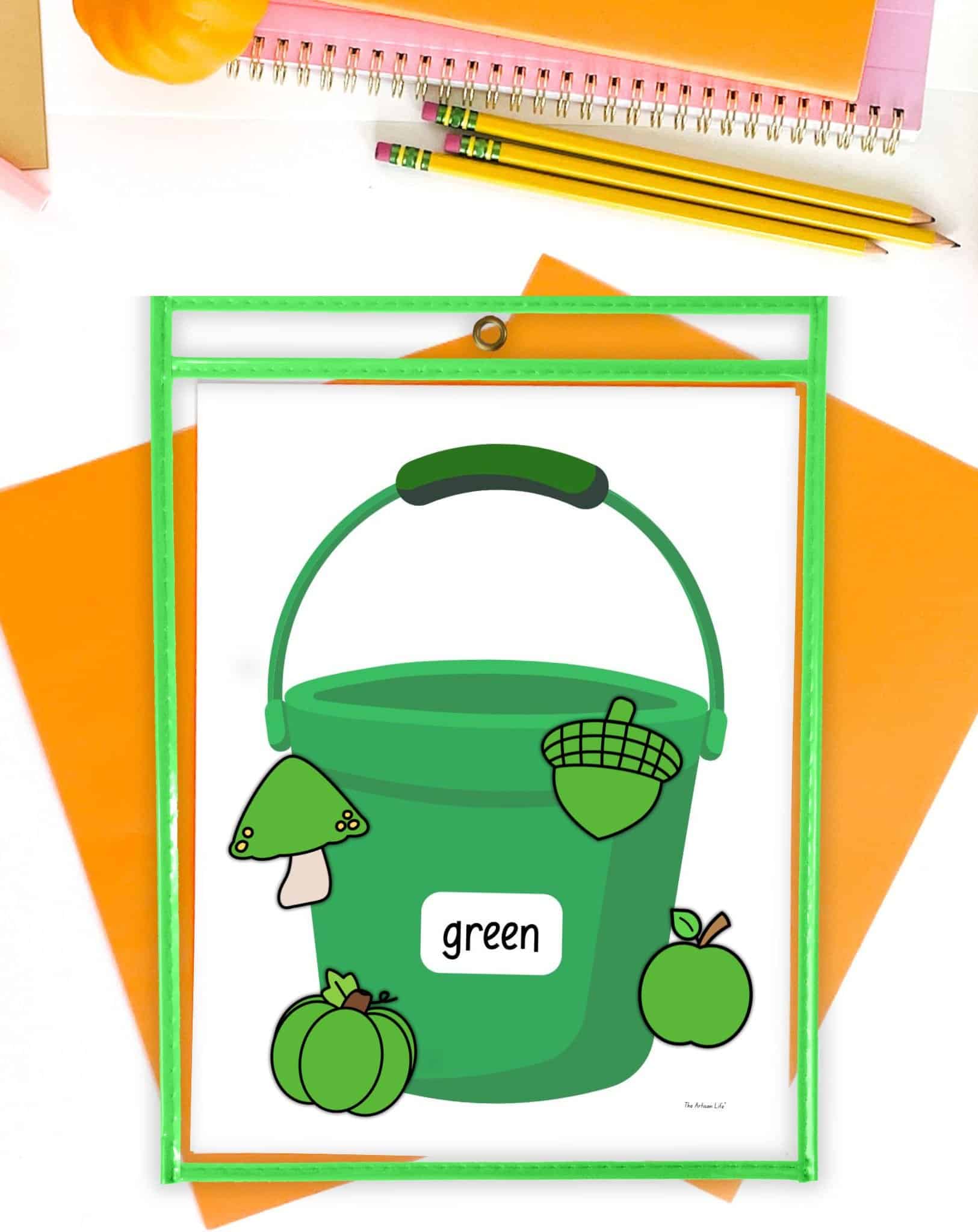 A printed green bucket in a green dry erase pocket with four green objects - a pumpkin, an apple, a mushroom, and an acorn. The items are on a desk with orange papers.