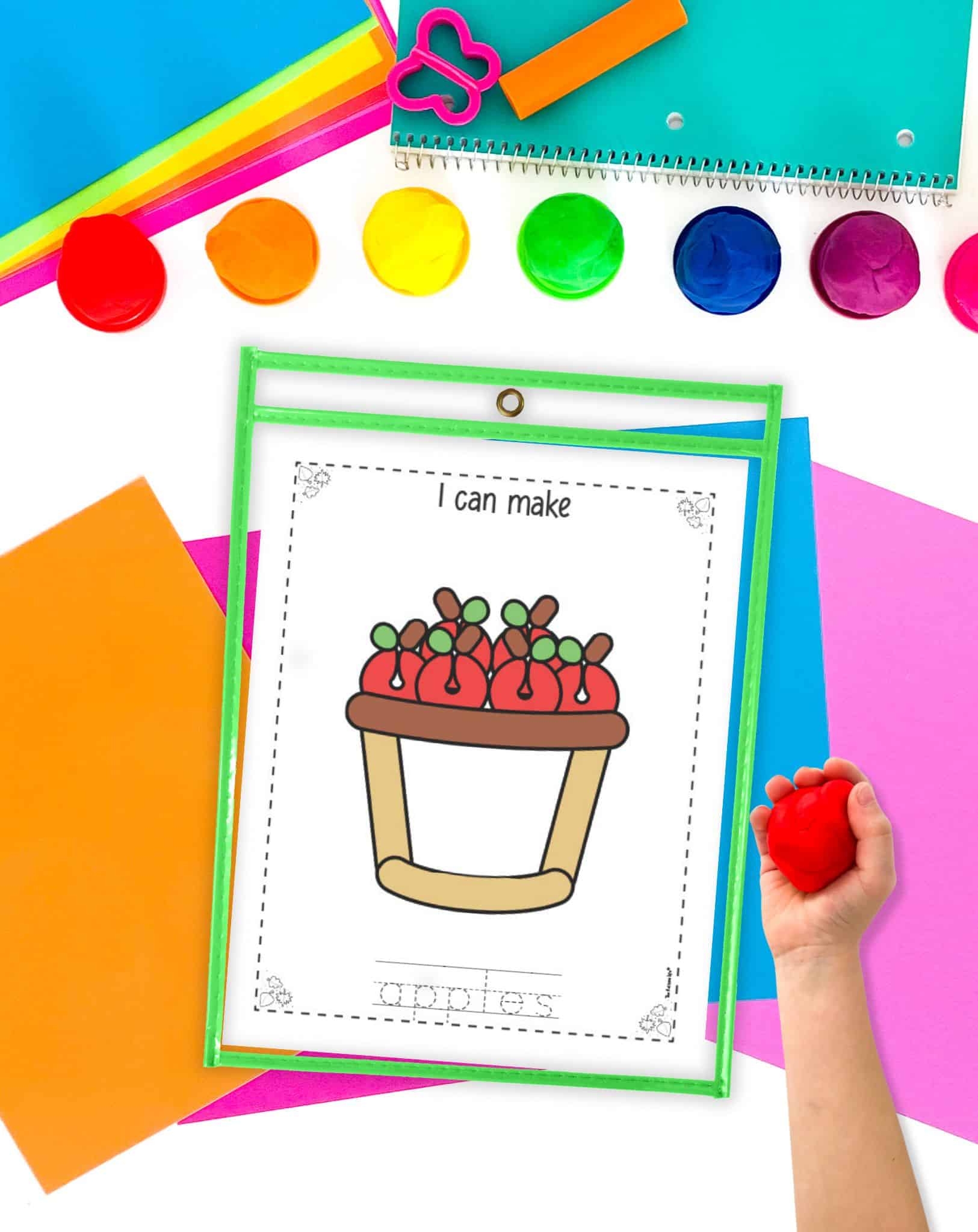 A basket of apples play dough mat in a dry erase pocket. A child's hand holding a ball of play dough is visible in the foreground