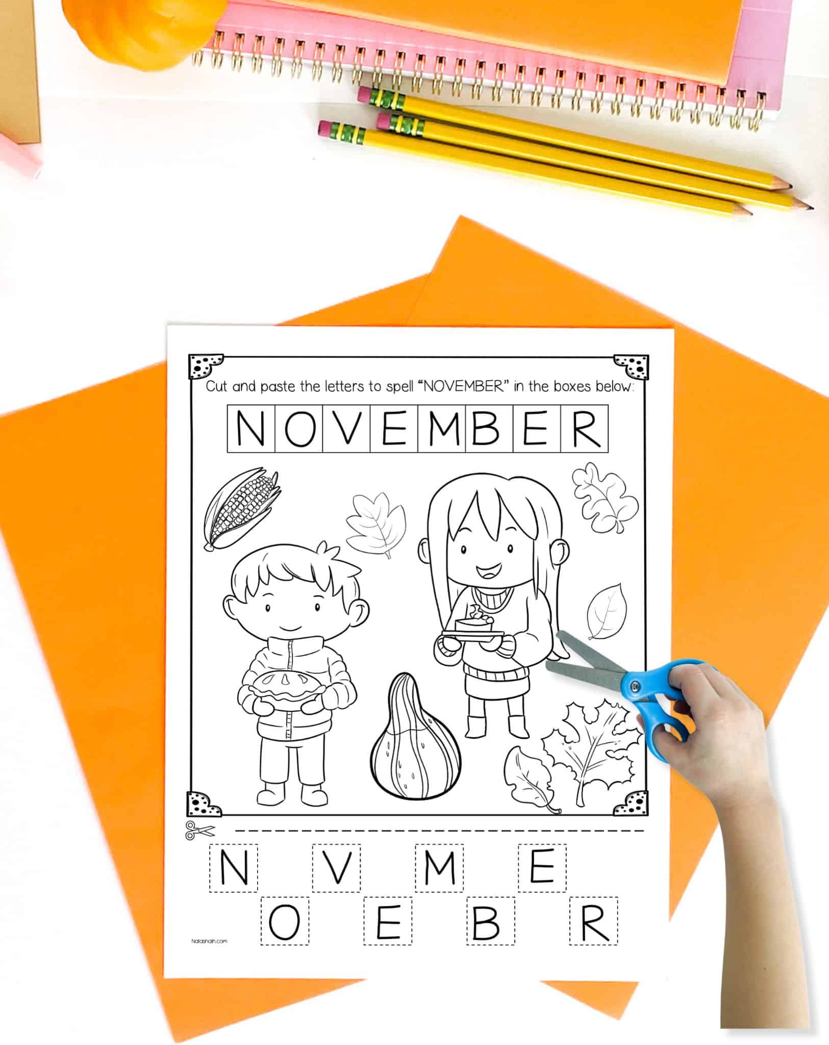 A November cut and paste worksheet is pictures to color and letter tiles to cut and paste. It is on a table with orange papers. A child's hand holding scissors is visible in the foreground.