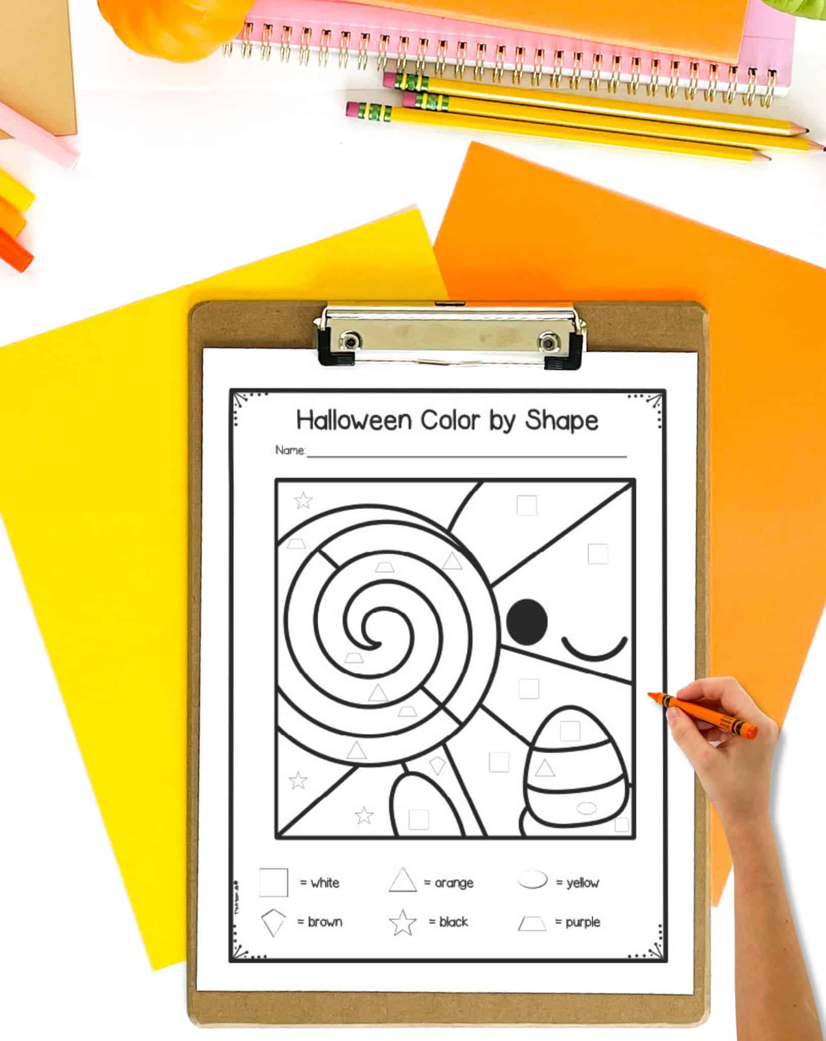 A cute Halloween ghost color by shape page on a clipboard. The clipboard is on a table with yellow and orange papers.