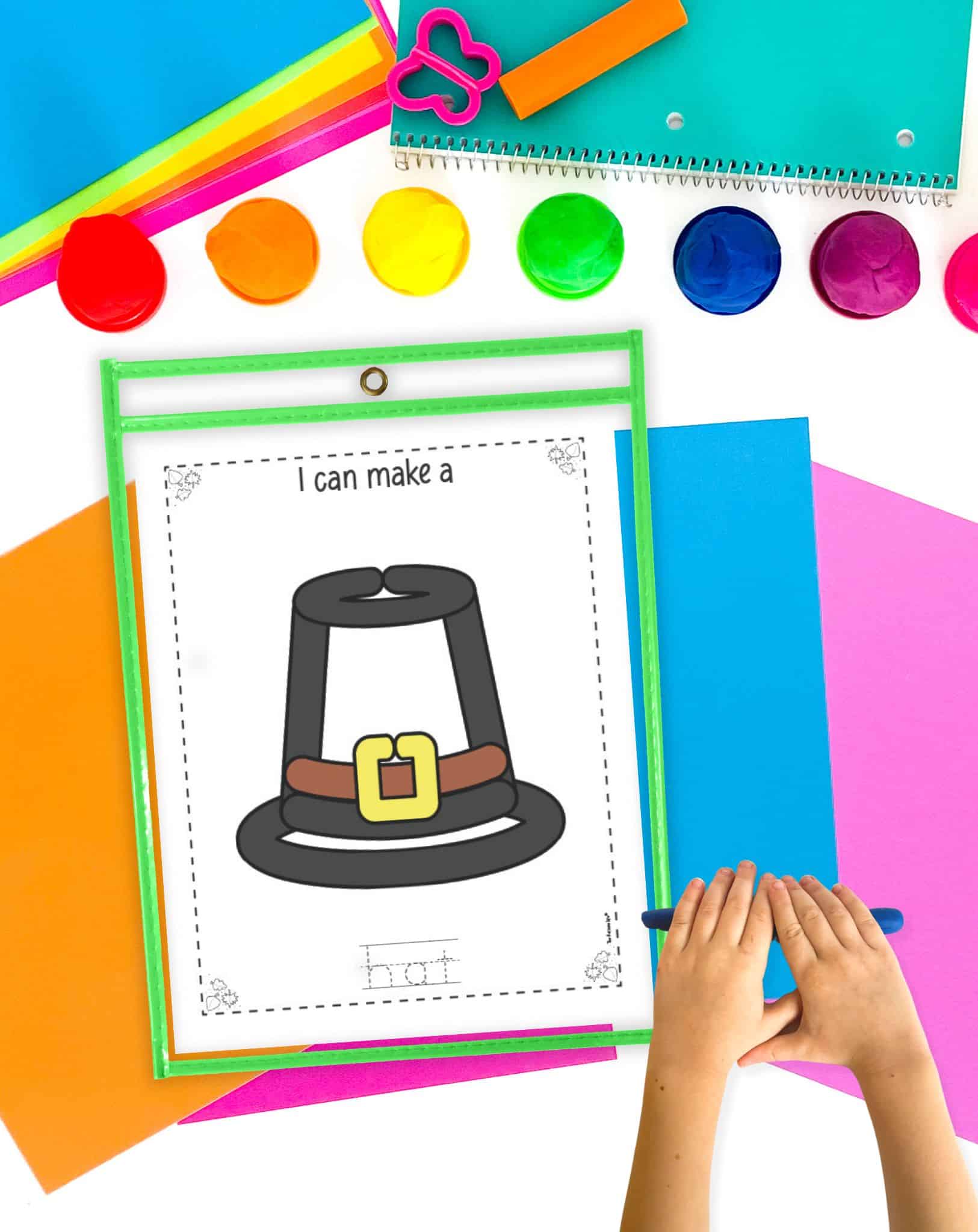 A Pilgrim hat play dough mat in a dry erase pocket on a table with colorful papers and play dough. A child's hands rolling out play dough are visible in the foreground