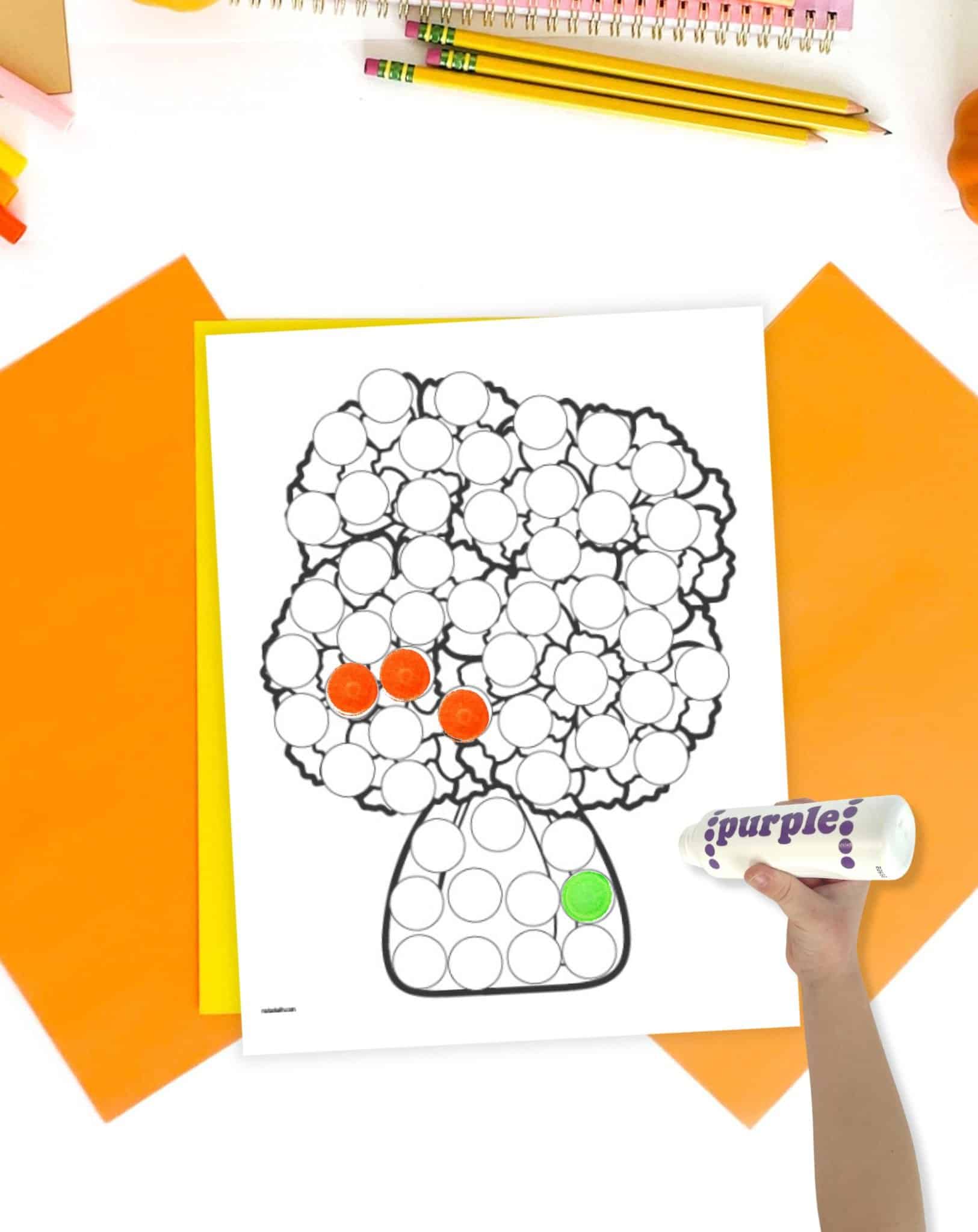 A day of the dead do marker page with flowers on a table with orange and yellow papers. A child's hand holding a dauber marker is visible in the foreground.