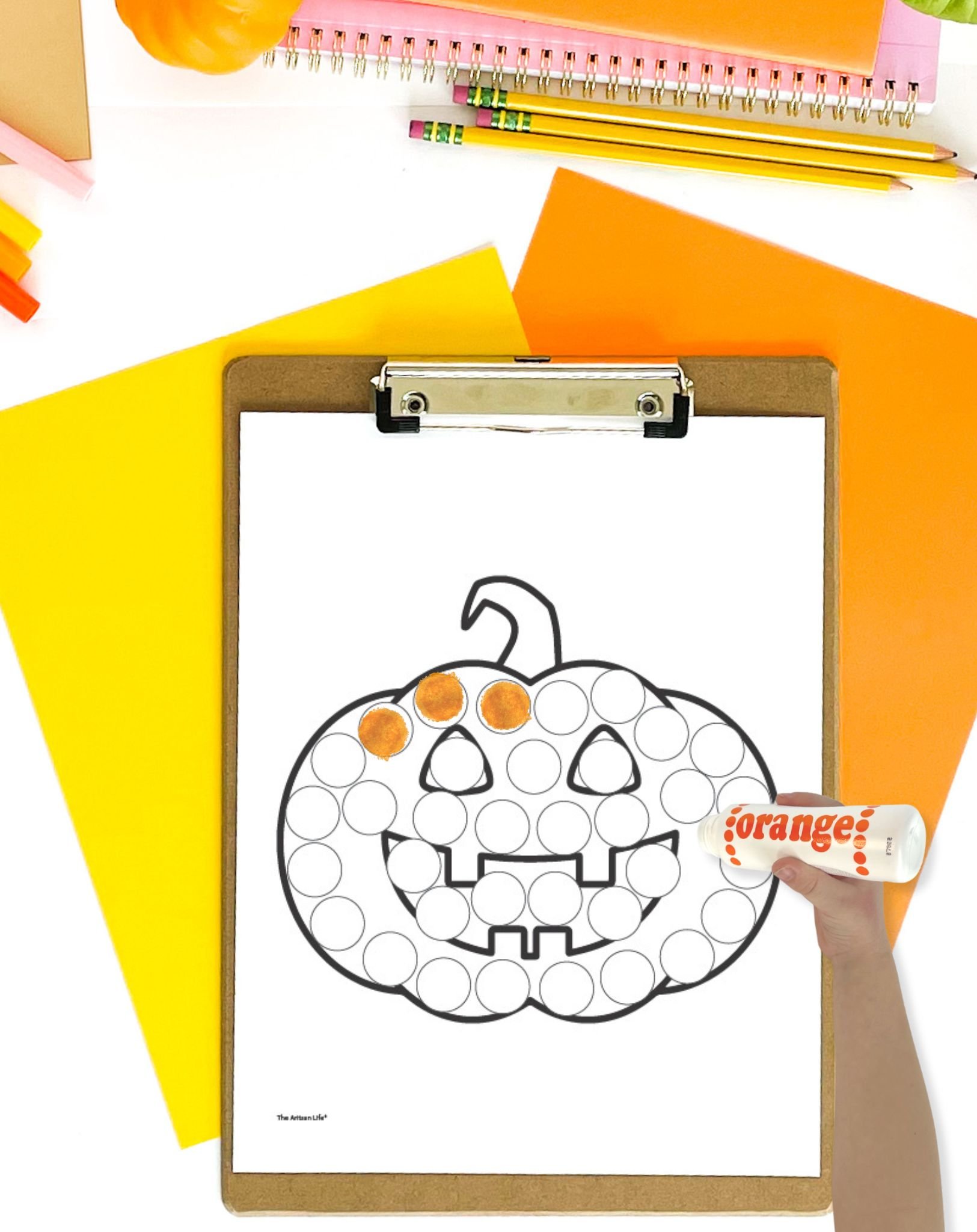 A Jack o'lantern dot marker page on a clip board with orange and yellow papers. A child's hand holding an orange dauber marker is visible in the foreground