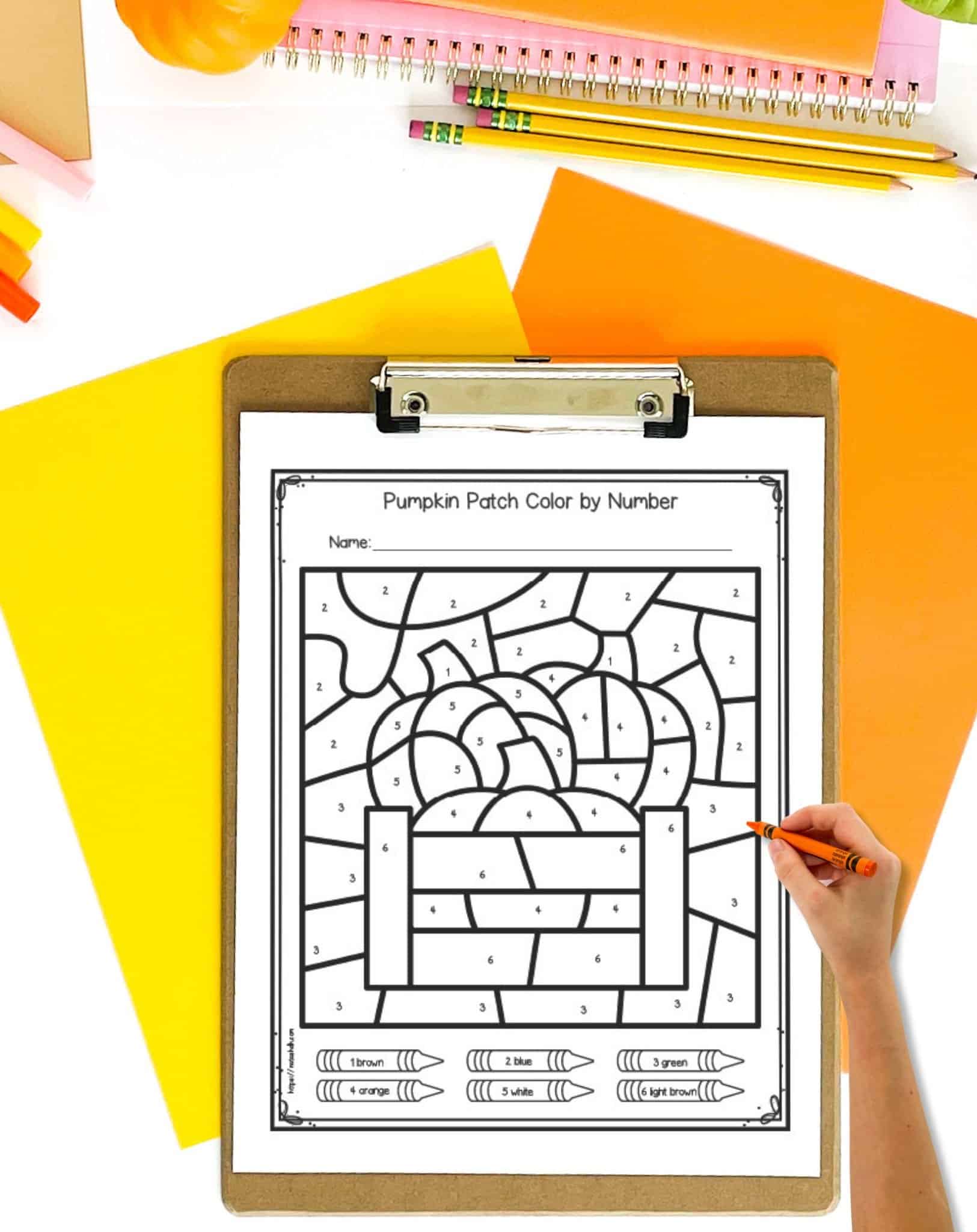 An un colored pumpkin themed color by number page on a clip board. The clip board is on a table with orange and yellow papers. A hand holding a crayon is visible in the foreground.