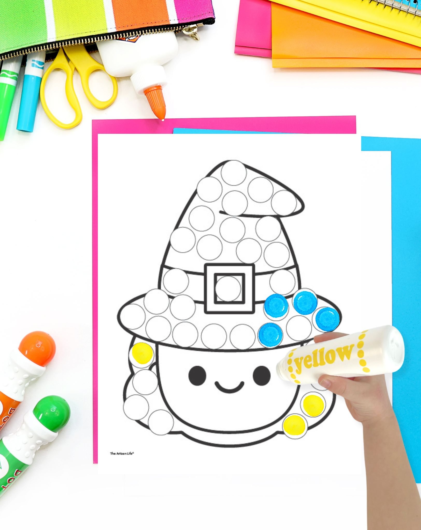 A cute witch face dot marker coloring page on a desk with colorful papers and dot markers