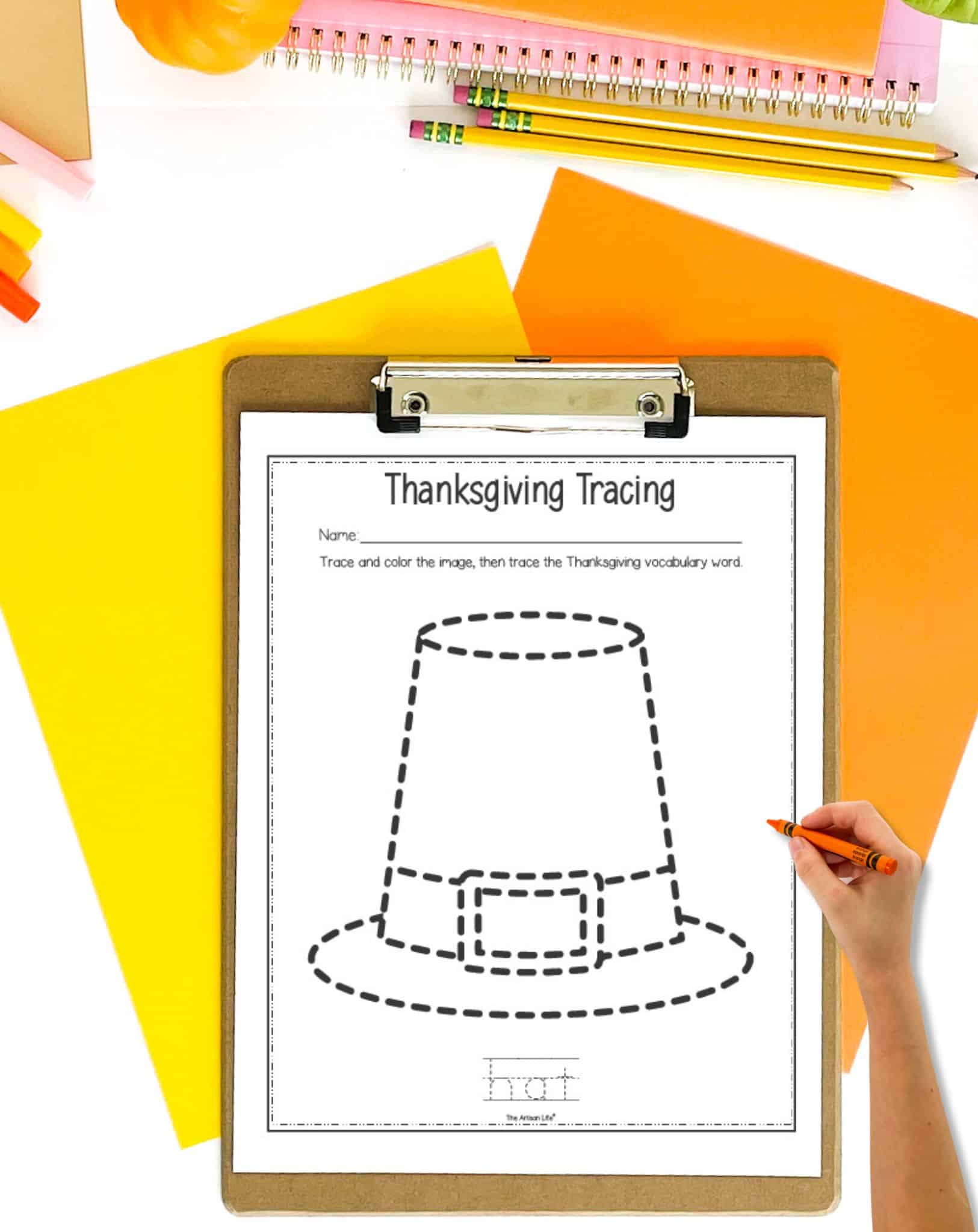 A pilgrim hat trace and color page on a clipboard. The clipboard is on a. table with orange and yellow papers. A child's hand holding a crayon is visible in the foreground.