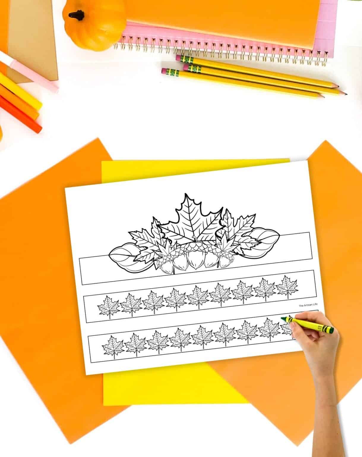 A fall headband craft print out on a table with orange papers and pencils. A child's hand holding a crayon is visible in the foreground