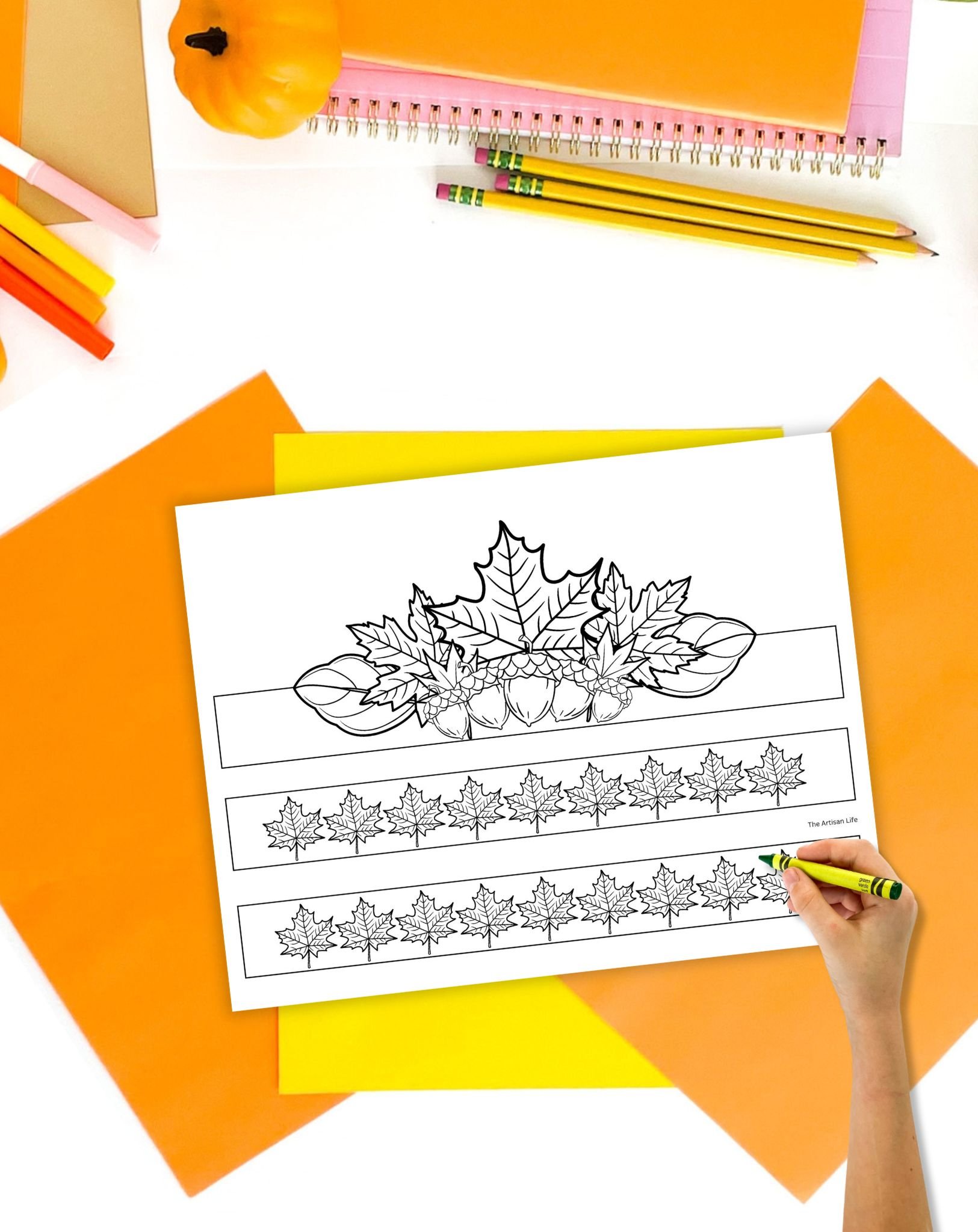 A fall headband craft print out on a table with orange papers and pencils. A child's hand holding a crayon is visible in the foreground