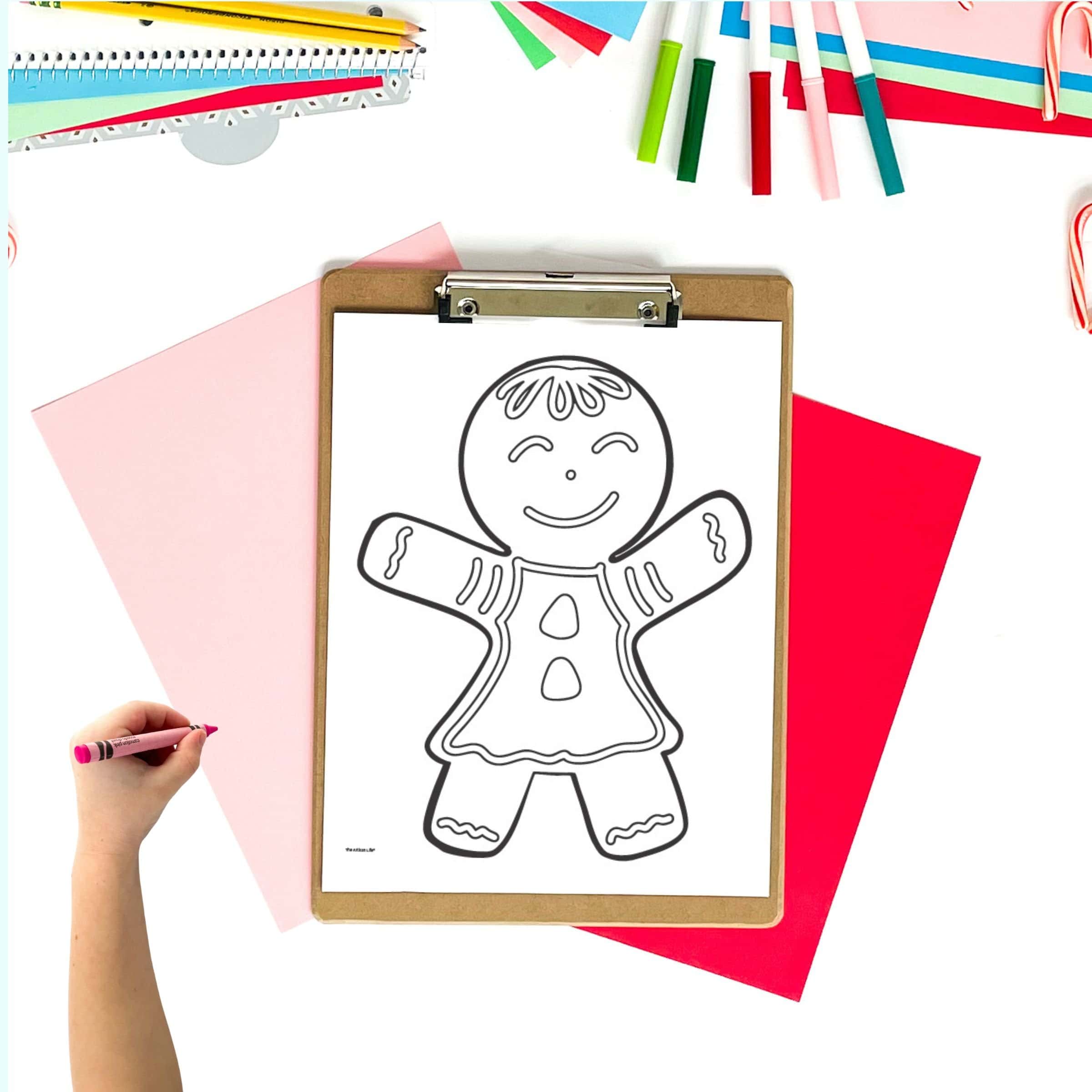 a gingerbread girl coloring page on a clipboard. The clipboard is on a desk with colorful Christmas paper supplies. A child's hand holding a crayon is visible in the foreground.