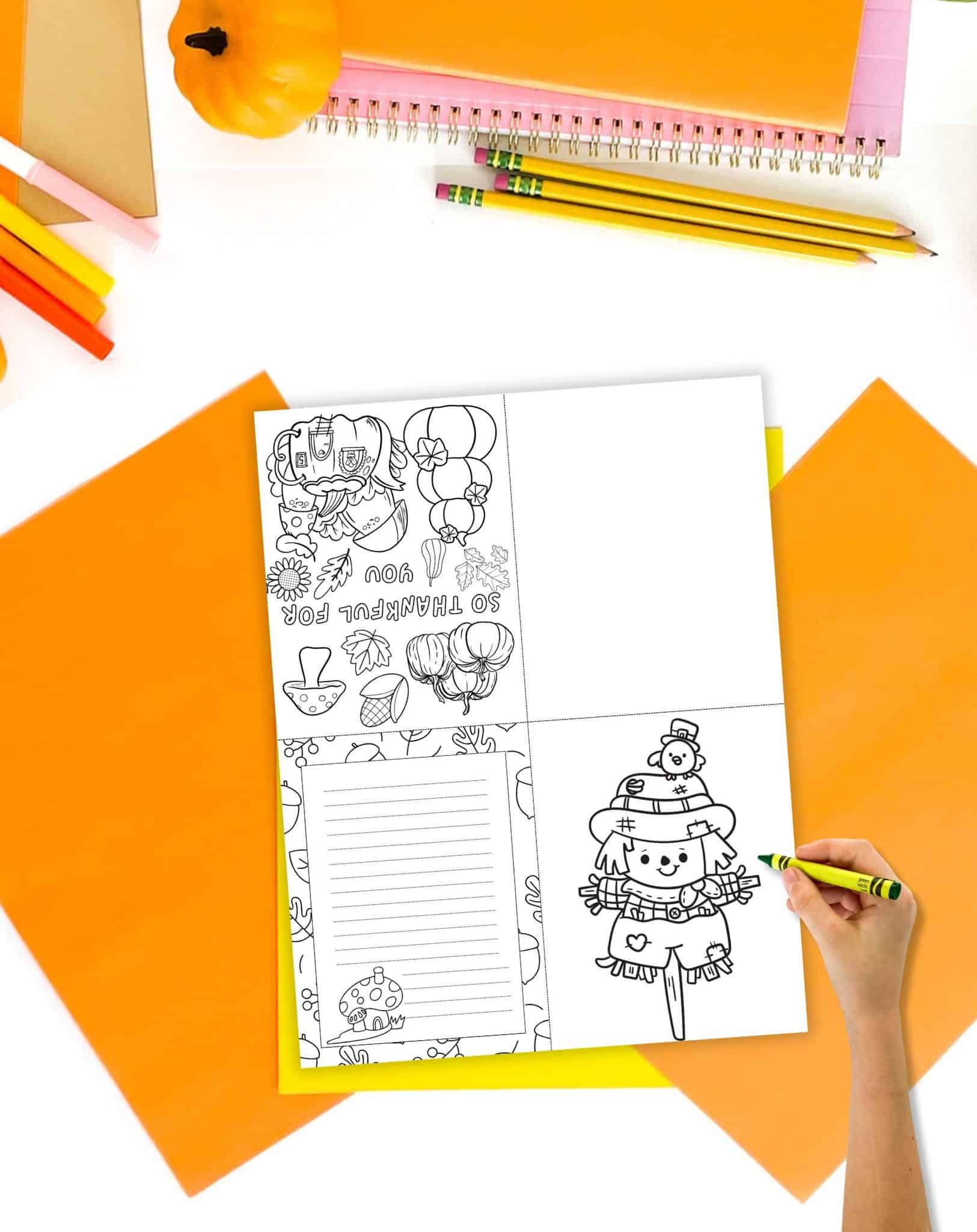 A Thanksgiving gratitude card showing a scarecrow, mushrooms, and pumpkins on a desk with orange papers and pencils. A child's hand holding a rayon is visible in the foreground