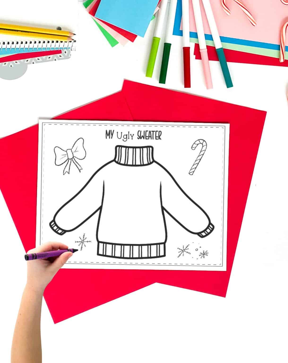 a design an ugly sweater page on a table with christmas papers. A child's hand holding a rayon is visible in the foreground.