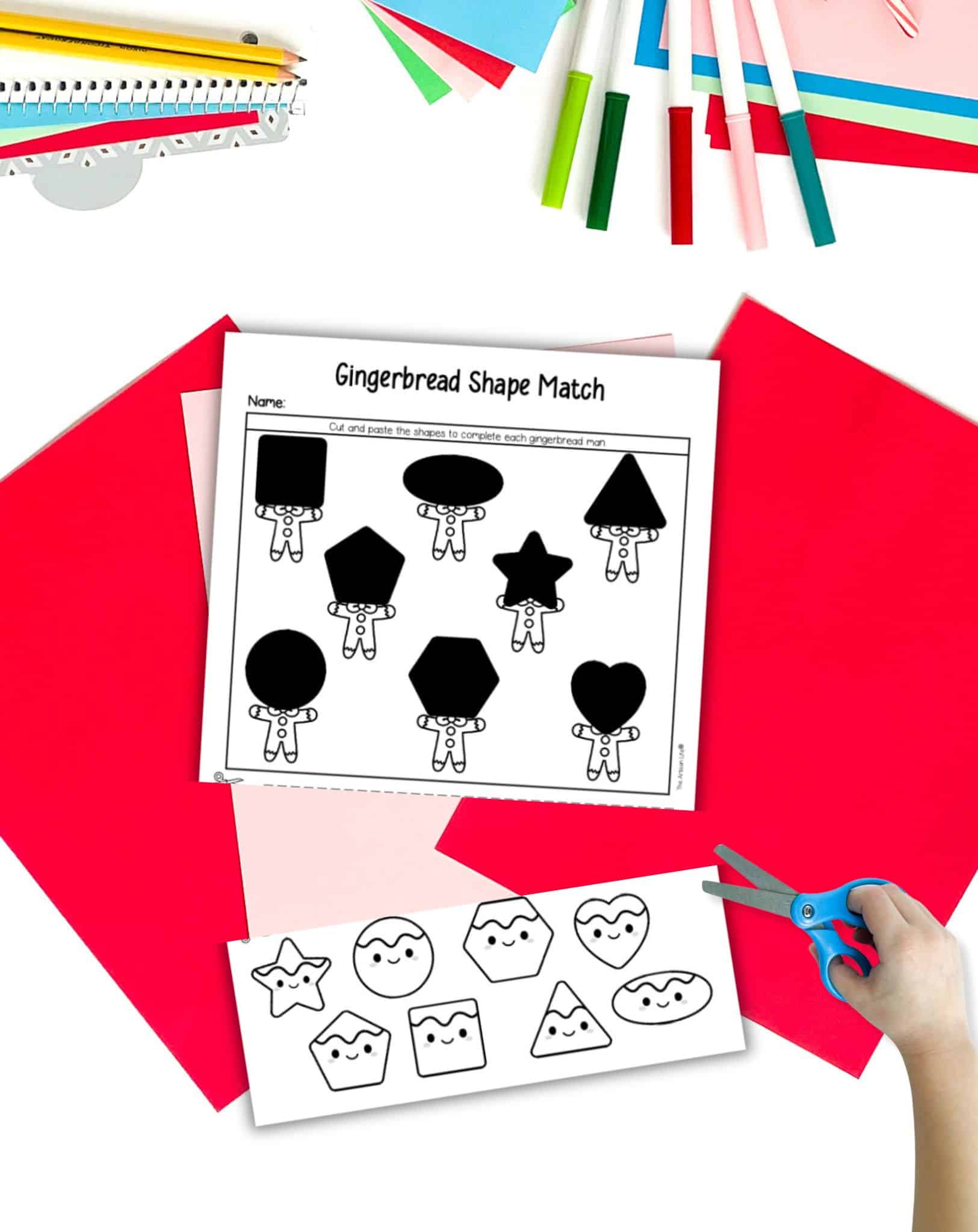 A gingerbread shape matching activity page that is cut along a dotted line. The page is on a table with Christmas stationary items. A child's hands holding scissors is visible in the foreground