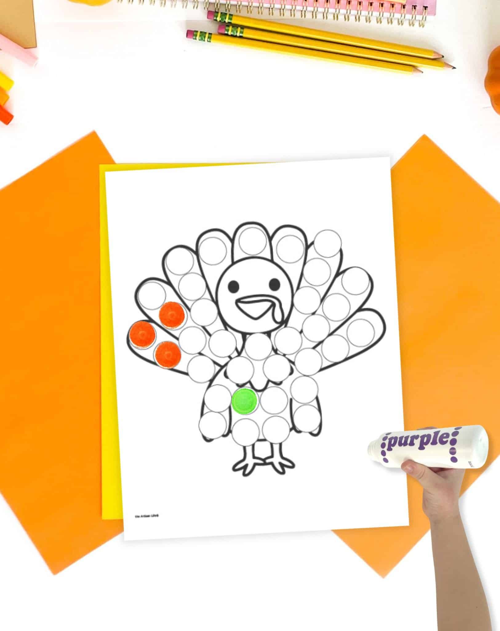 A turkey themed dot marker coloring page on a table with orange and yellow papers. A child's hand holding a dot marker is visible in the foreground. 