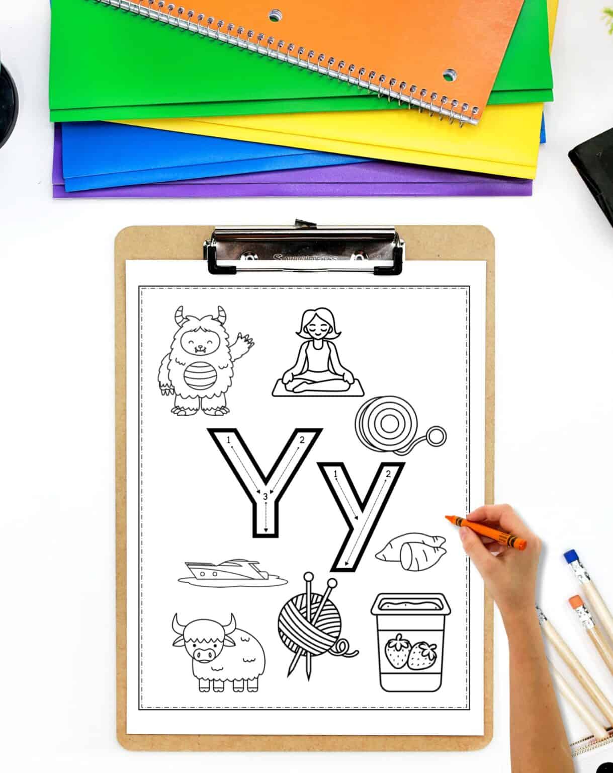 A letter y coloring page on a clipboard. A child's hand holding a crayon is visible in the foreground.