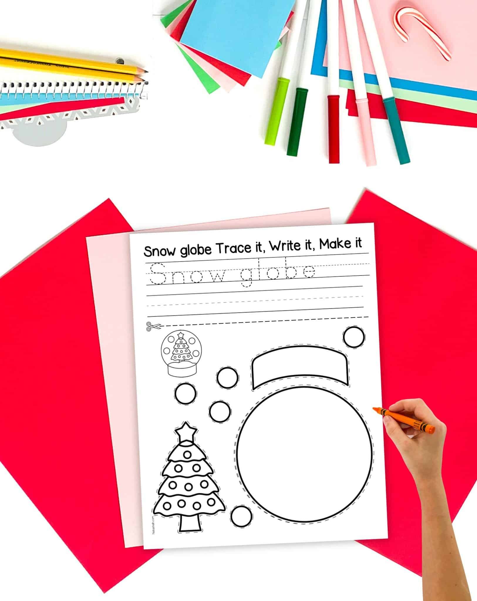 a black and white snow globe cut and paste craft on a table with Christmas stationary items. A child's hand holding a crayon is visible in the foreground 