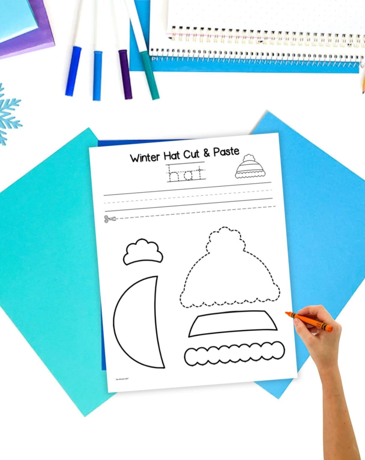 a black and white cut and paste craft for a winter that. It is on a table with blue stationary items. A child's hand holding a crayon is visible in the foreground