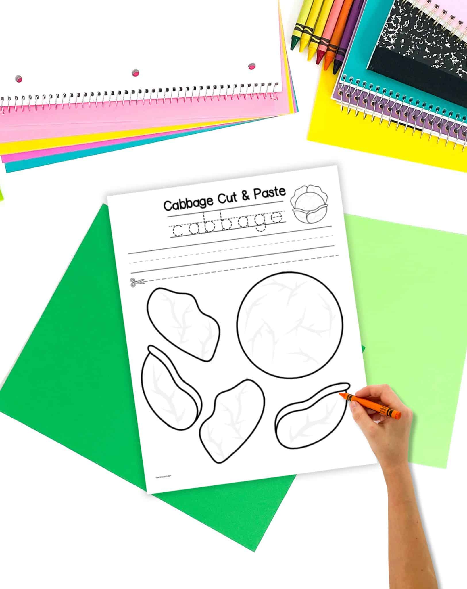 A black and white cabbage cut and paste craft on a table with green papers. A child's hand holding a crayon is visible in the foreground.