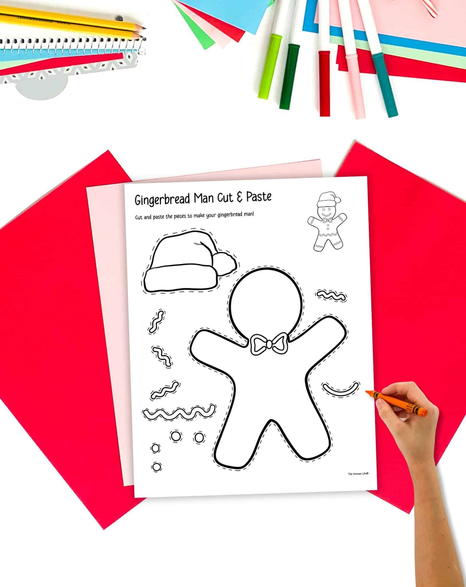 a gingerbread man cut and paste craft print out on a table with colorful Christmas stationary items. A child's hand is holding a pencil in the foreground