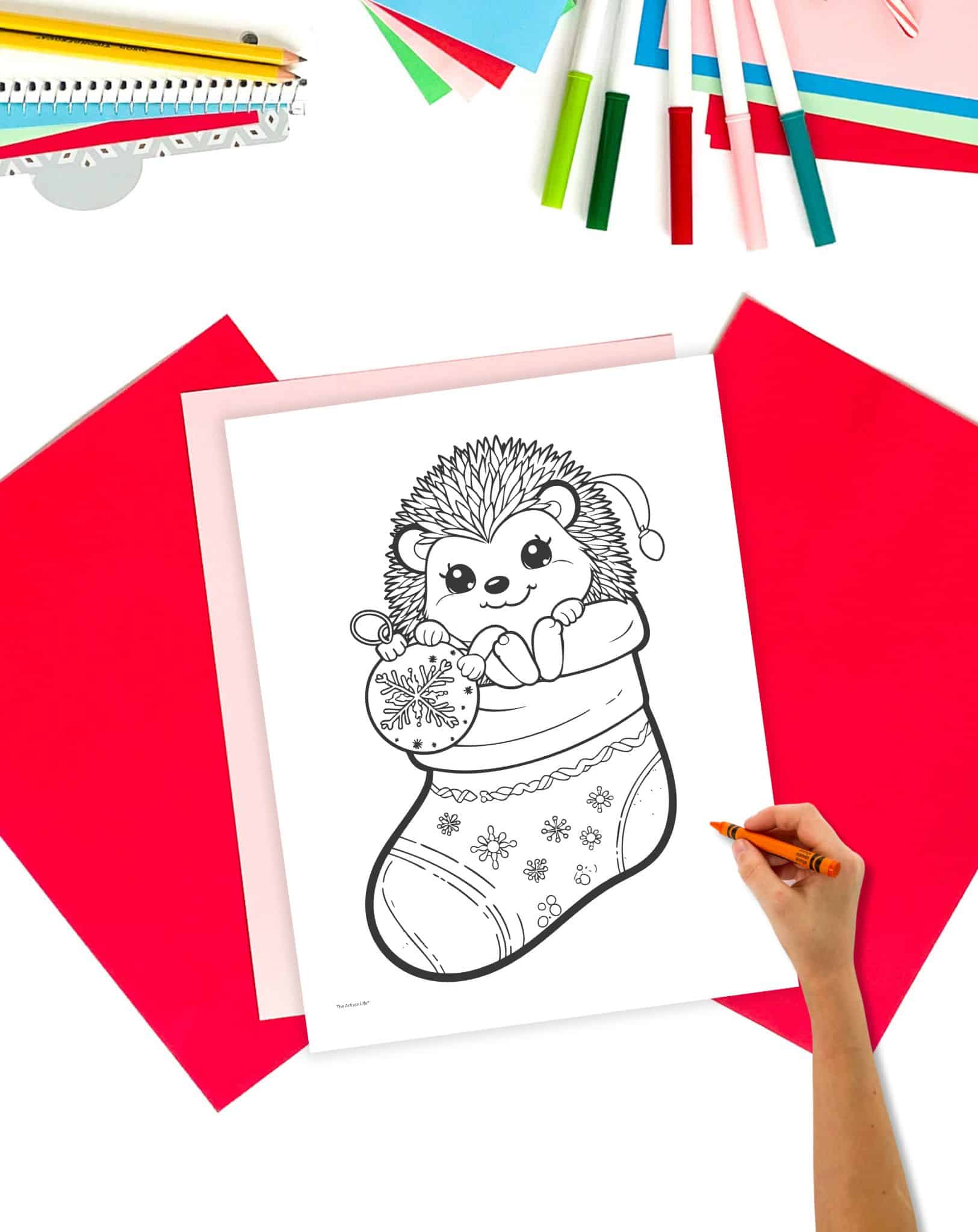 a coloring page with a cute hedgehog in a christmas stocking. the page is on a table with christmas stationary. A child's hand holding a crayon is visible in the foreground