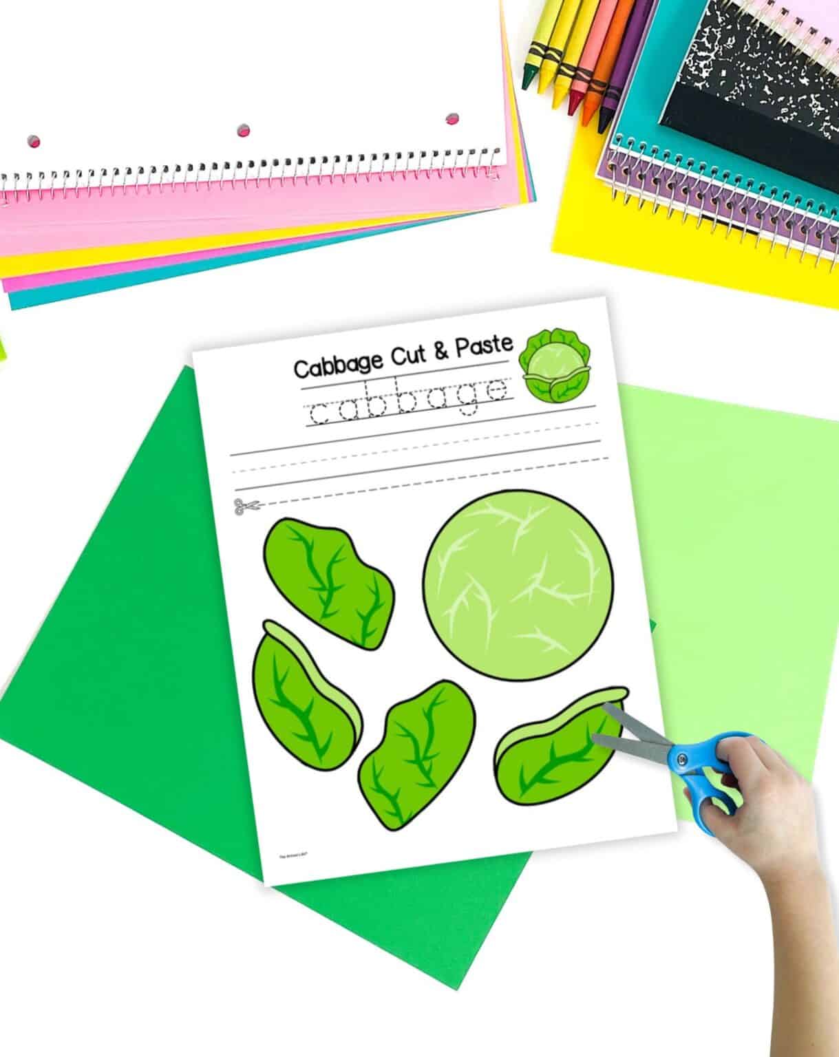 A cabbage cut and paste craft on a table with green papers. A child's hand holding scissors is visible in the foreground.
