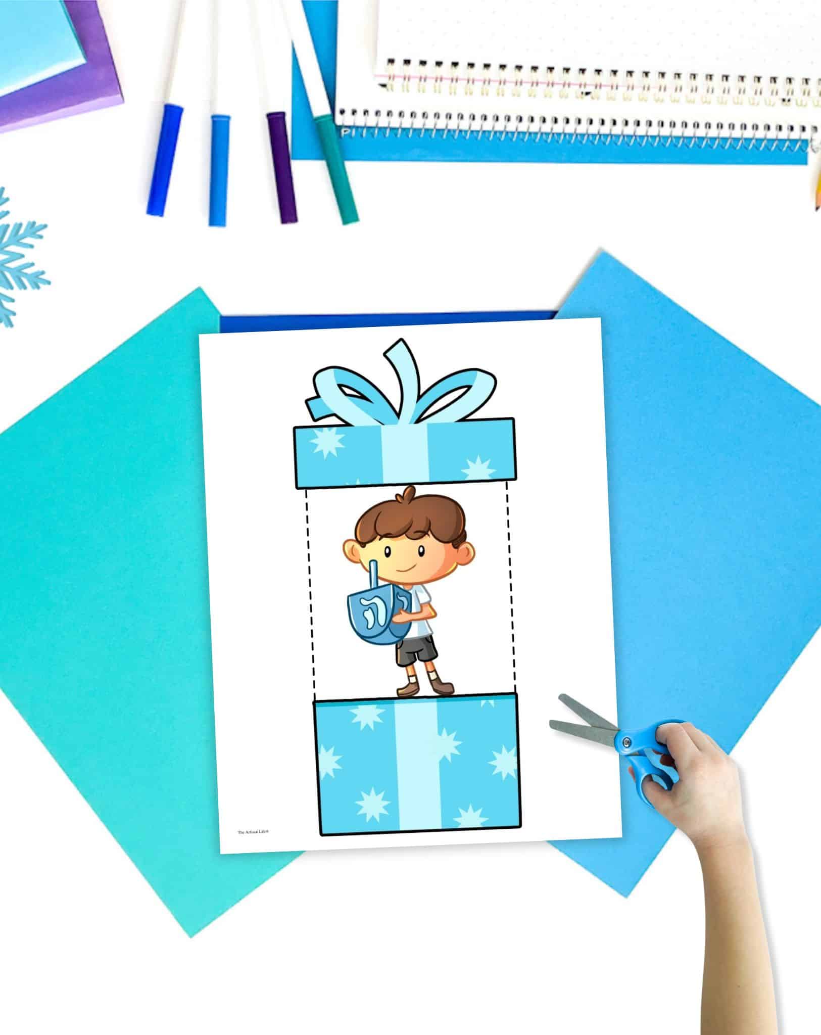 A printable Hanukkah card showing a boy holding a dredge. it is on a table with blue stationary items.