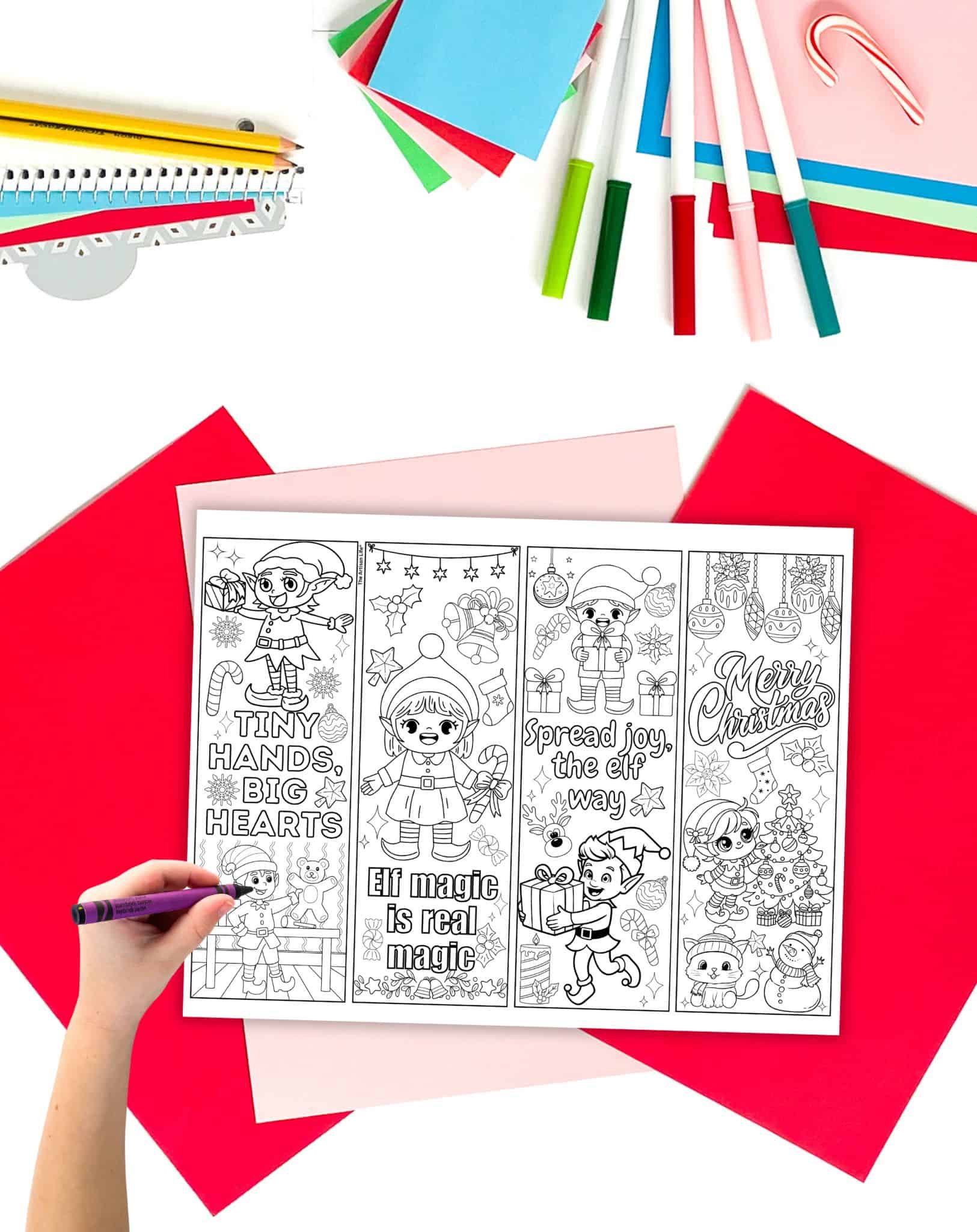 A page of elf coloring page bookmarks on a table with Christmas stationary. A child's hand holding a crayon is visible in the foreground