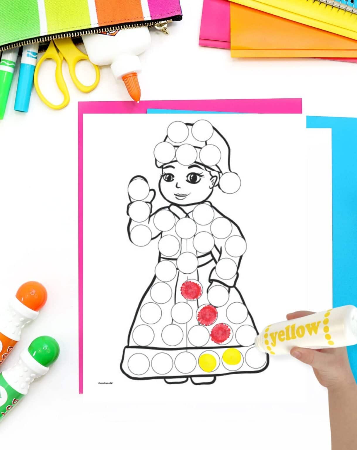 a partially completed mrs Claus dot marker page on a table with colorful marker and papers. A child's hadn't holding a dot marker is visible in the foreground