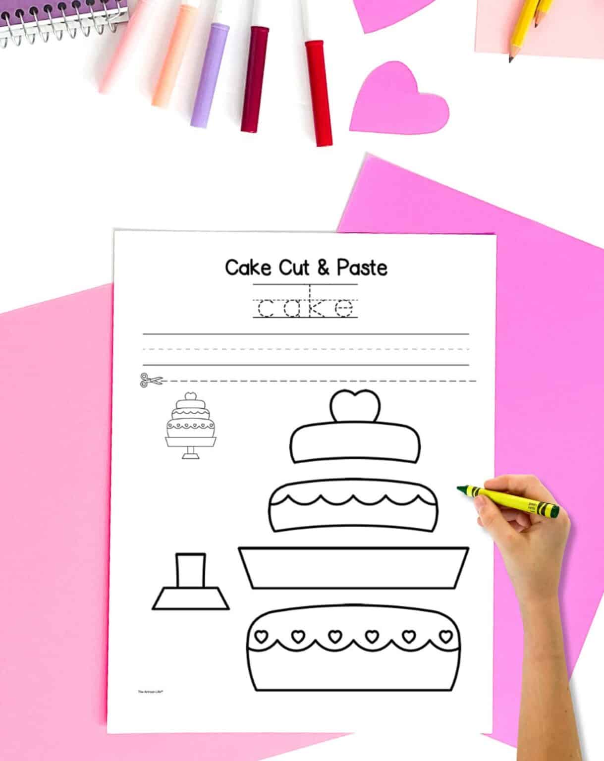 A cut and paste cake craft on a table with pink papers and markers. A child's hand holds a crayon in the foreground.