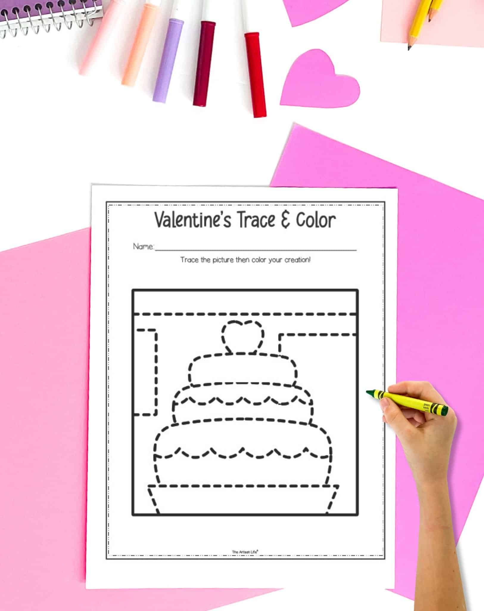 a Valentine's Day trace and color worksheet on a table with pink papers. A child's hand holding a crayon is visible in the foreground