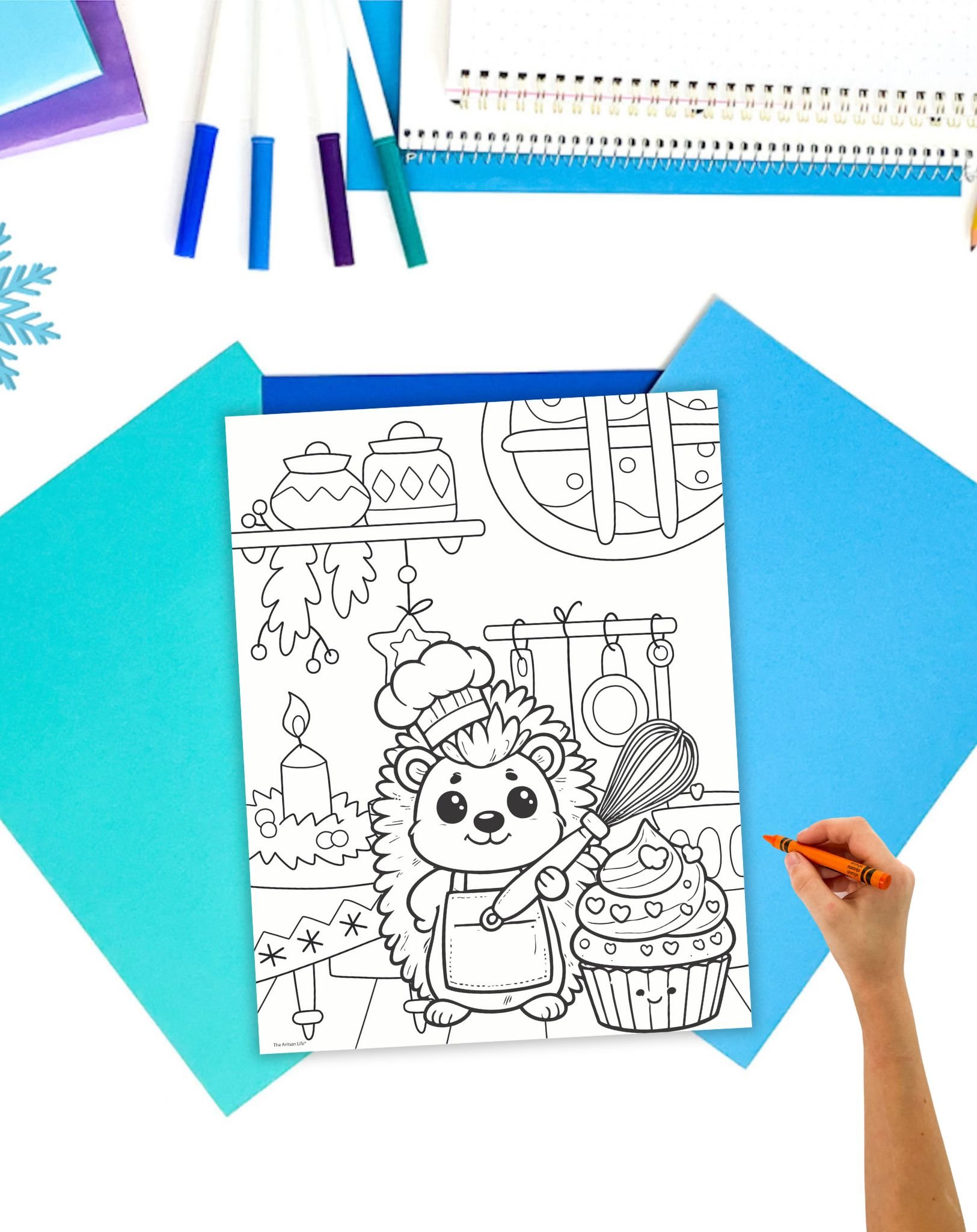 a coloring page showing a hedgehog with a cupcake in a kitchen decorated for winter. The page is on a desk with blue papers.