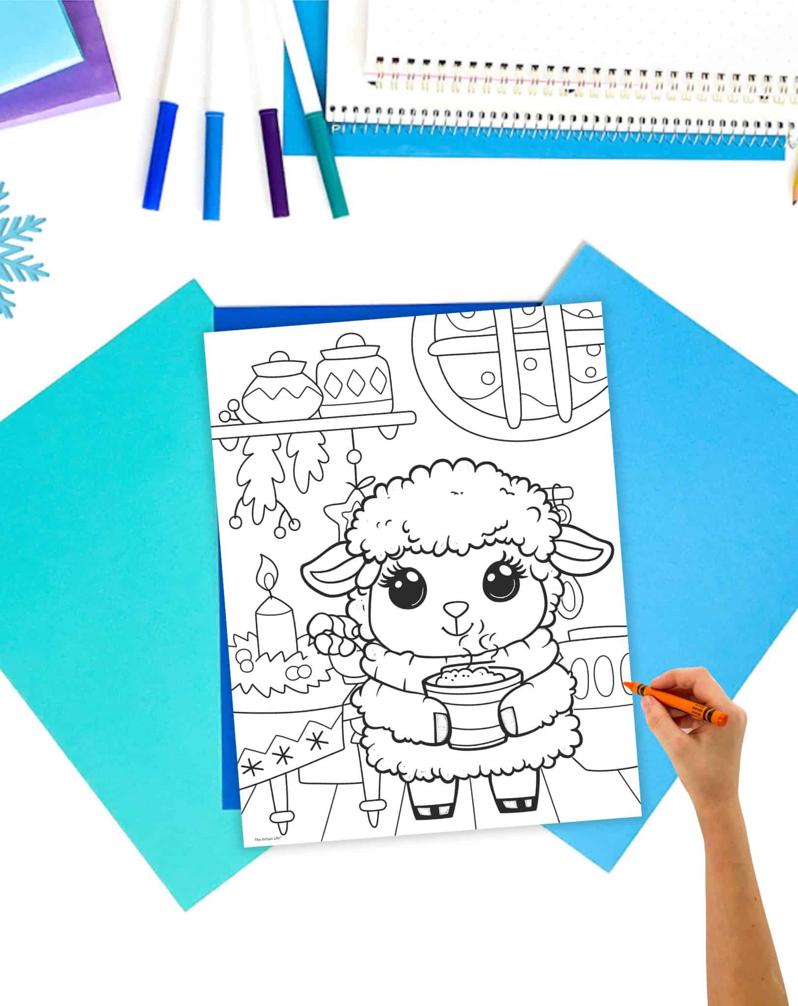 a coloring page of a cute winter lamb holding a steaming mug in a kitchen decorated for winter. The page is on a table with blue papers. A child's hand holding a crayon is visible in the foreground.