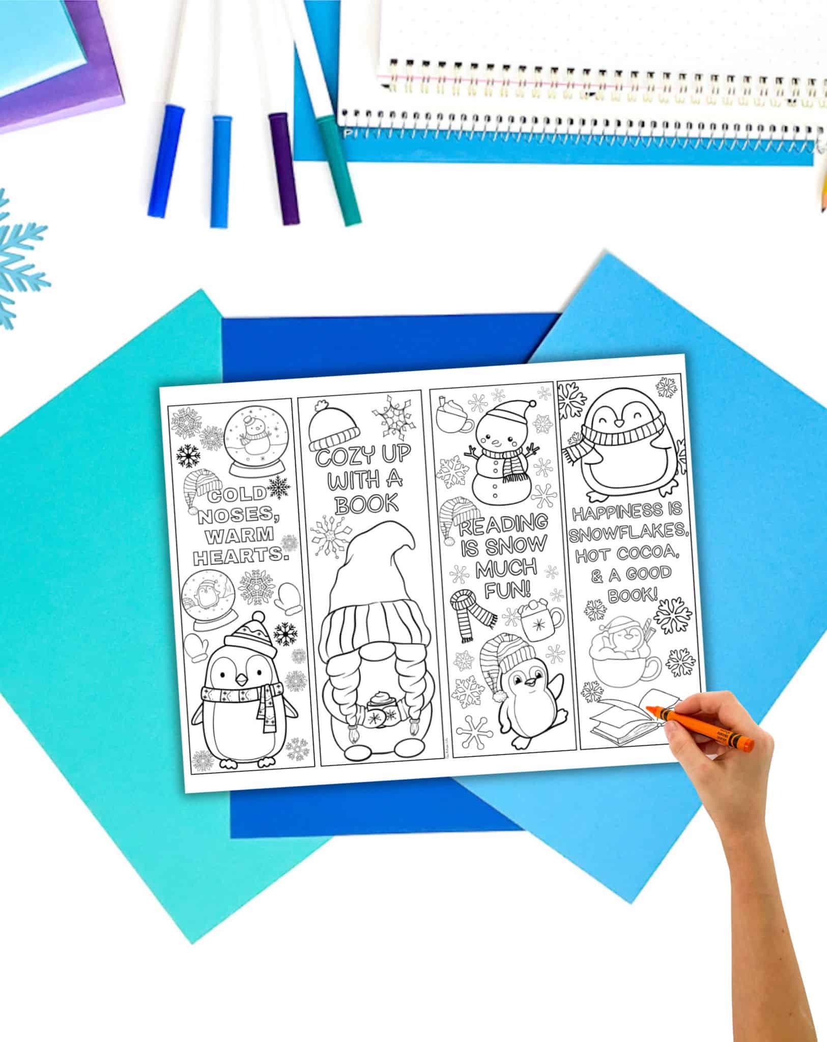 a page with winter bookmarks to color on a student desk with blue papers. A child's and holding a crayon is visible in the foreground