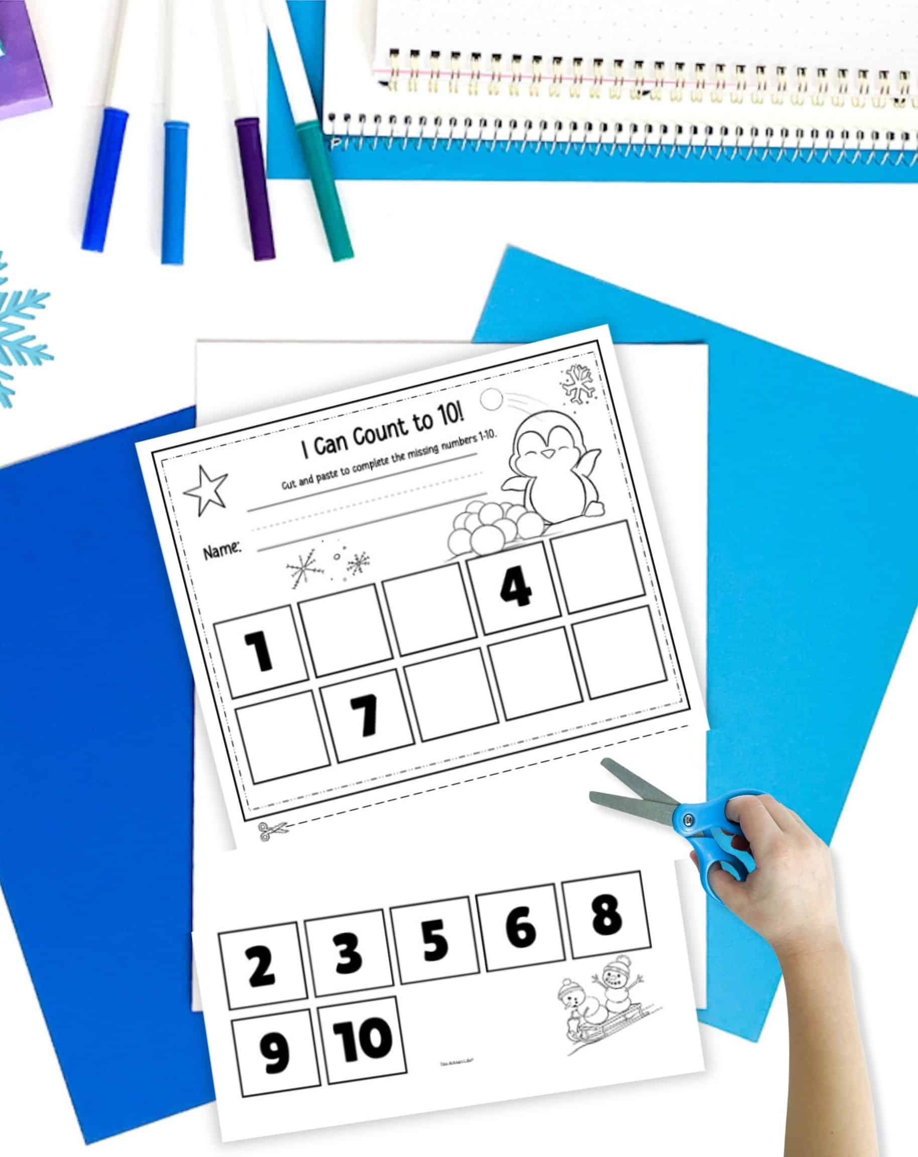 a cut in half winter cut and paste missing numbers worksheet. It is on a desk with blue papers. A child's hand is holding scissors in the foreground