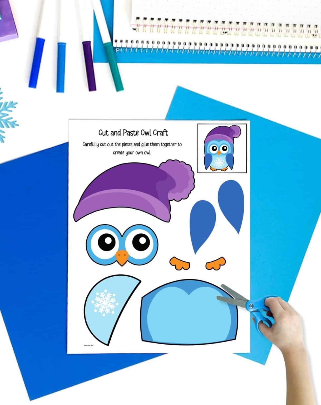 a winter owl cut and paste craft on a table with blue papers and markers. A child's hand holding scissors is visible in the foreground