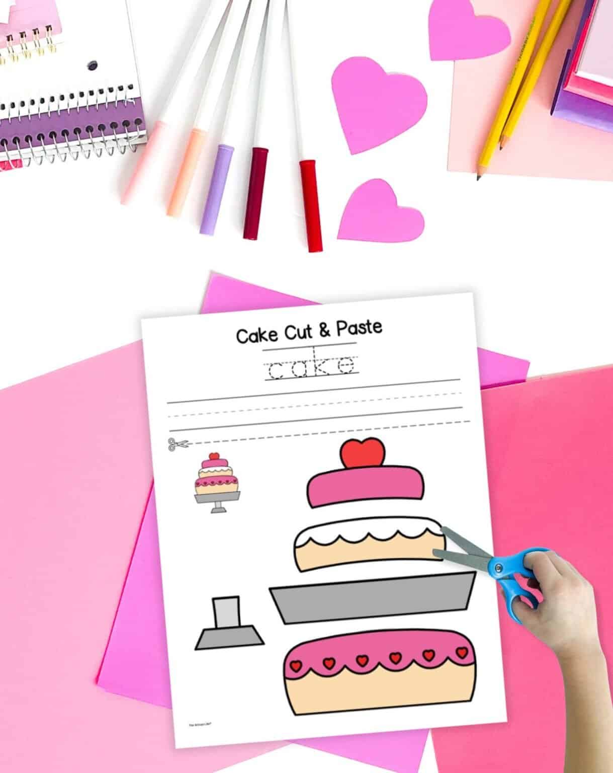A cut and paste cake craft on a table with pink papers and markers. A child's hand holds a pair of scissors in the foreground.